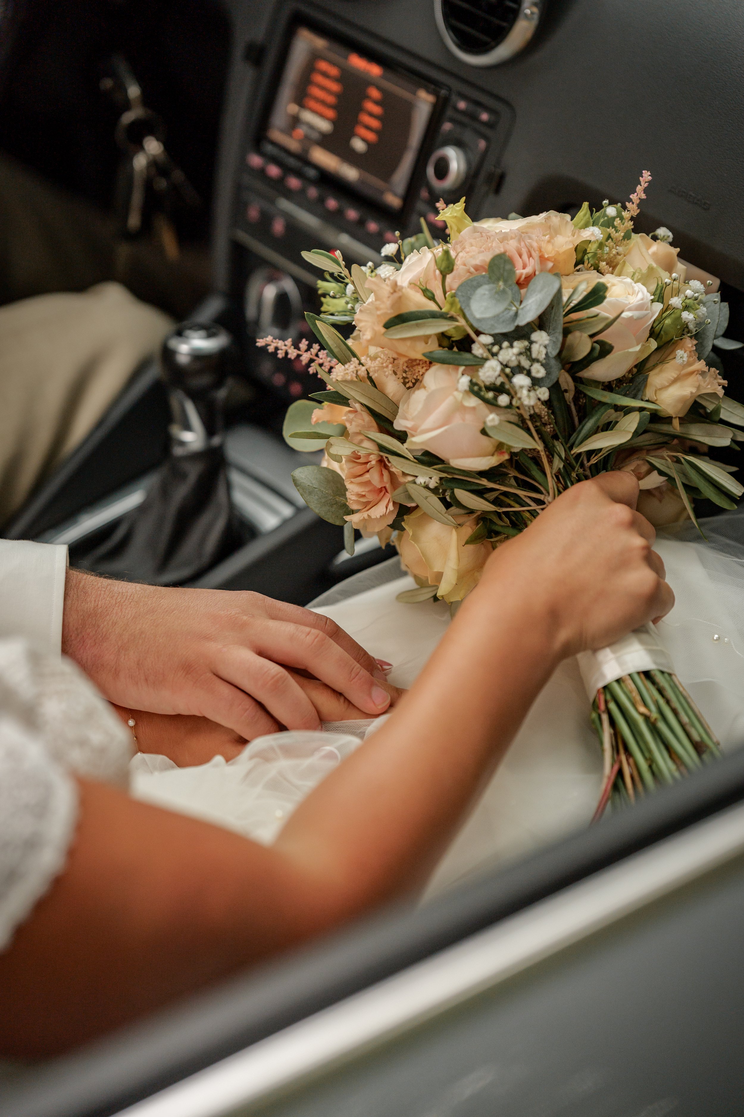 Een bruid in een witte jurk houdt een boeket met bloemen en groen in de auto, terwijl haar hand op de hand van de bruidegom rust. Gefotografeerd door Mateo Media.