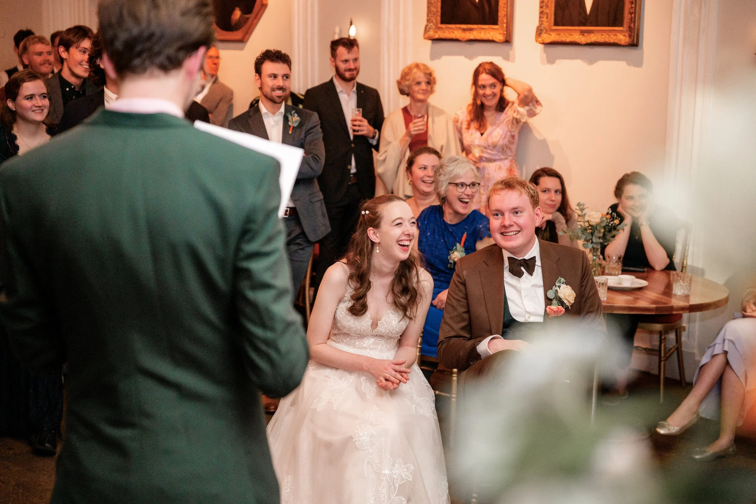 Een bruid en bruidegom lachen tijdens hun trouwceremonie, omringd door lachende gasten in een feestzaal. Gefotografeerd door professionele fotograaf Mateo Media.