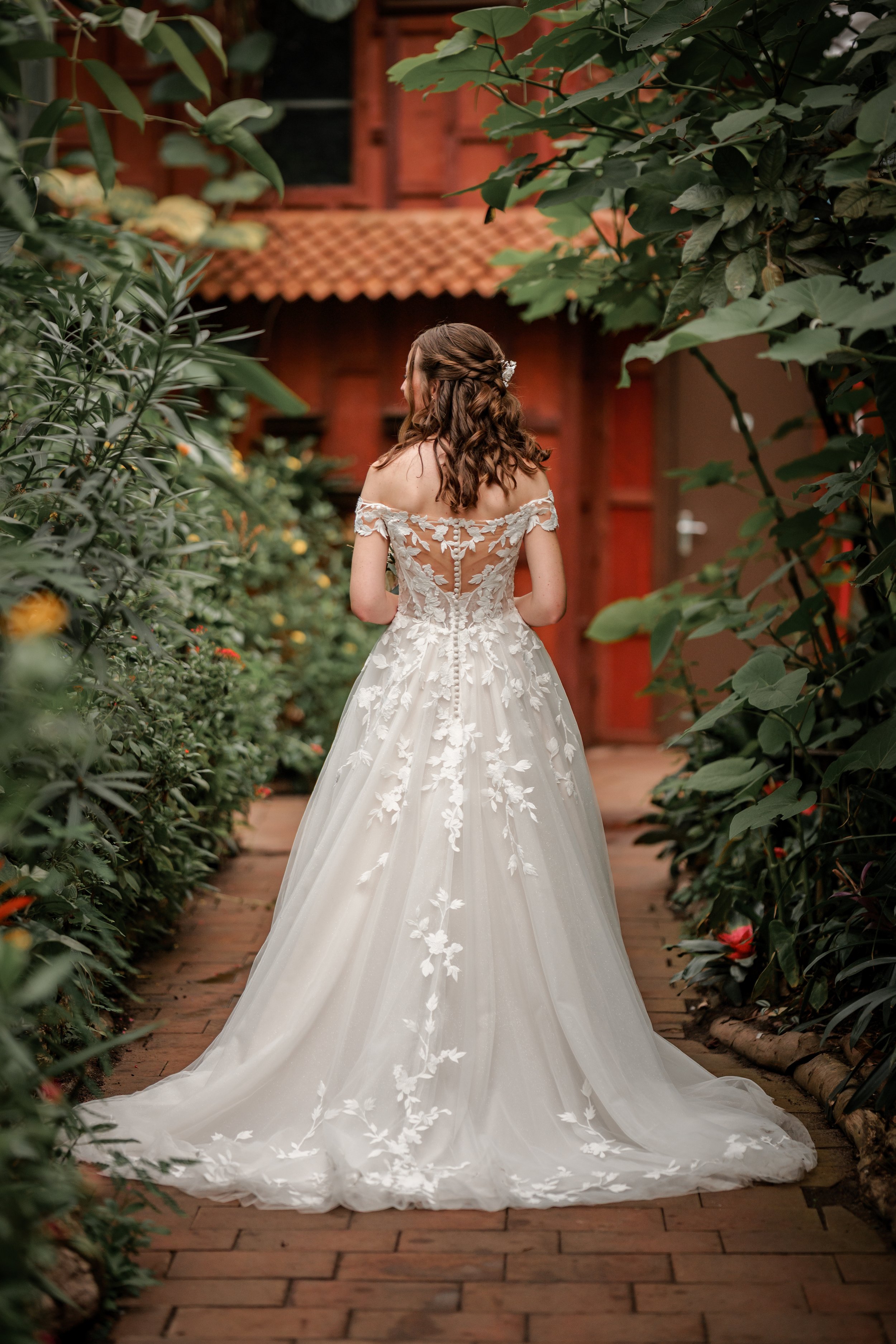 Een bruid in een witte, romantische trouwjurk met bloemen appliqué, staand op een stenen pad in een tuin met groene planten en bloemen. Vintage foto door fotograaf uit Gelderland.