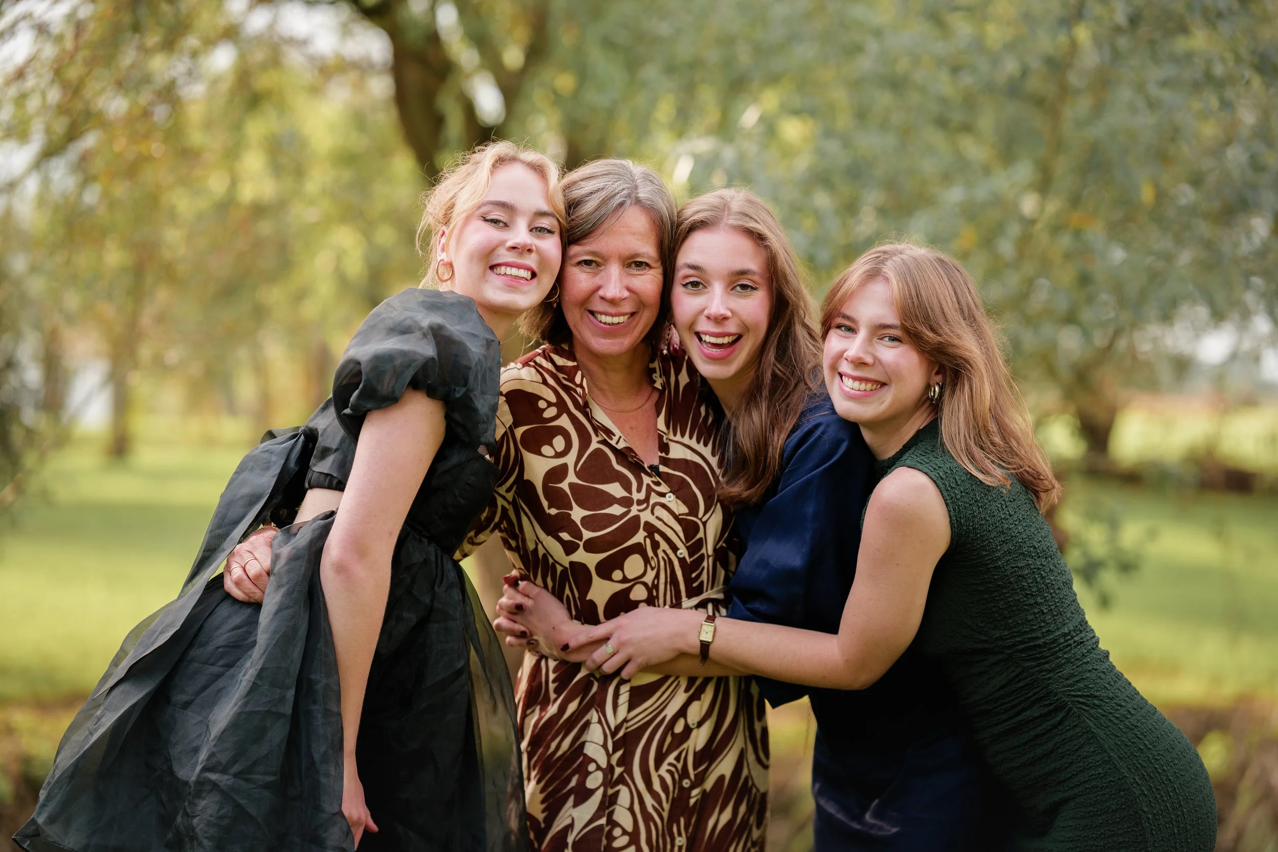 Vrouw samen met drie dochters die lachend in een park met groene bomen staan. Gezinshoot door professionele fotograaf uit Nederland.