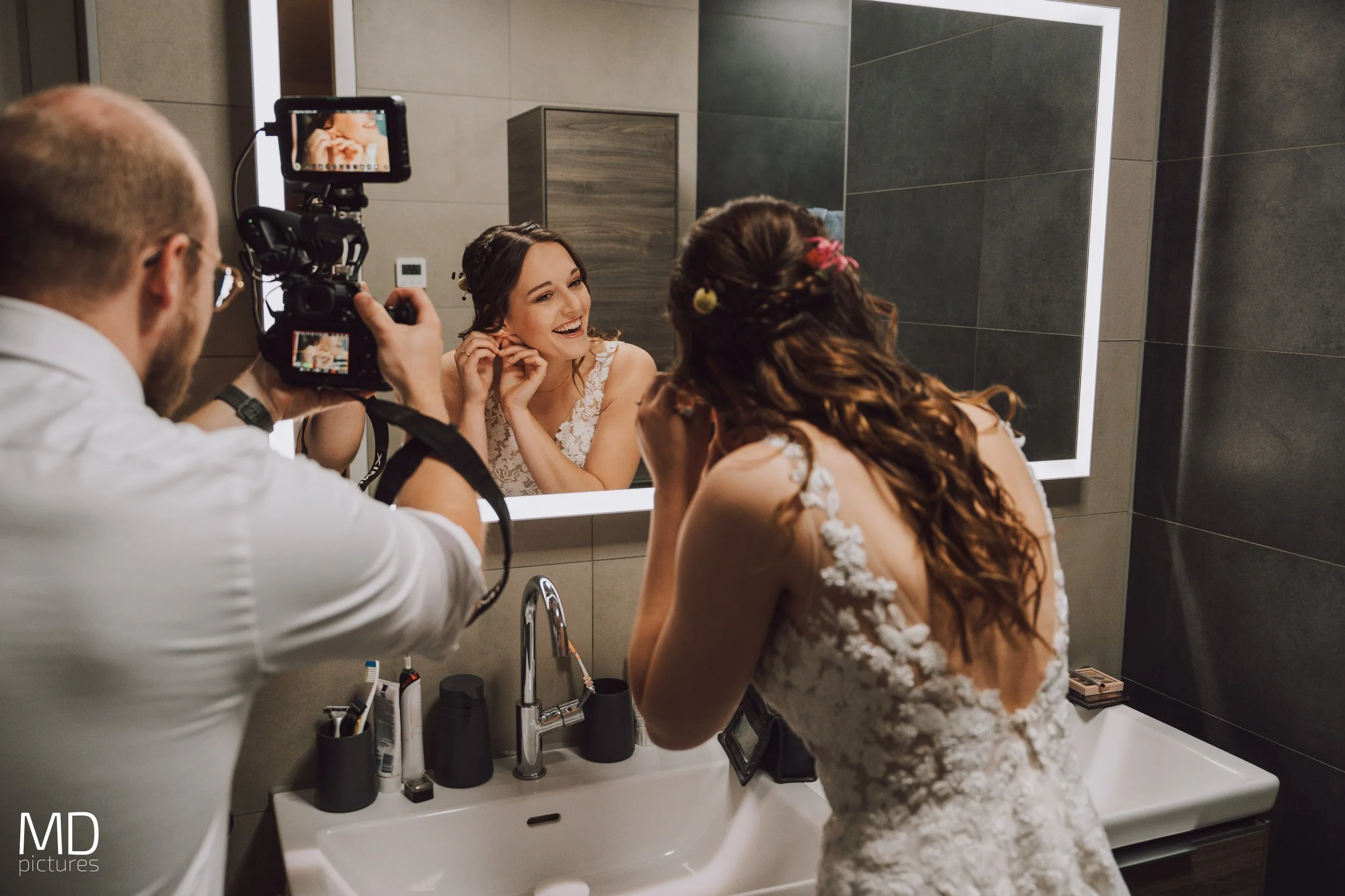 Een vrouw in een witte bruiloftsjurk die zichzelf in de spiegel opmaakt terwijl een fotograaf haar fotografeert in een badkamer.