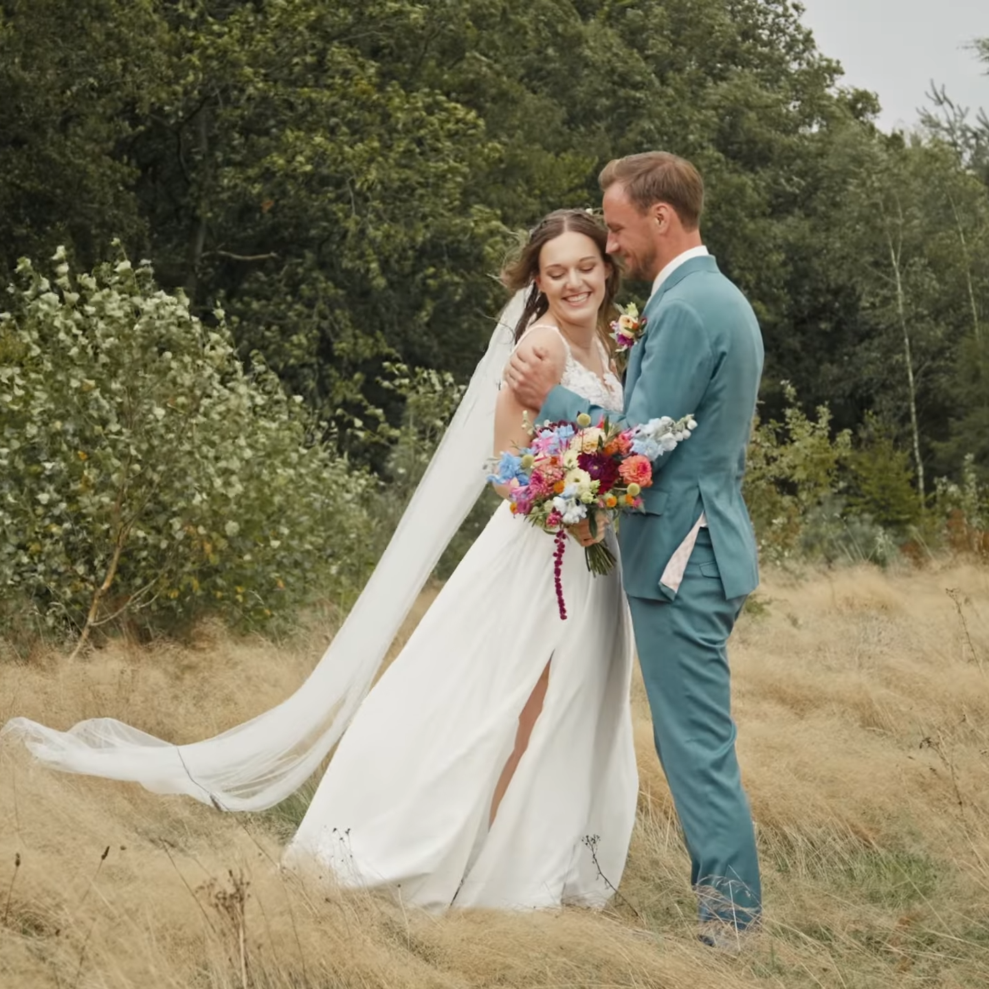 Twee mensen in een trouwjurk en pak staan samen in een veld met gras, omringd door bomen, lachen en kijken naar elkaar, de vrouw houdt een kleurrijk bloemenboeket vast. Shot uit film door professionele trouwvideograaf uit Nederland.