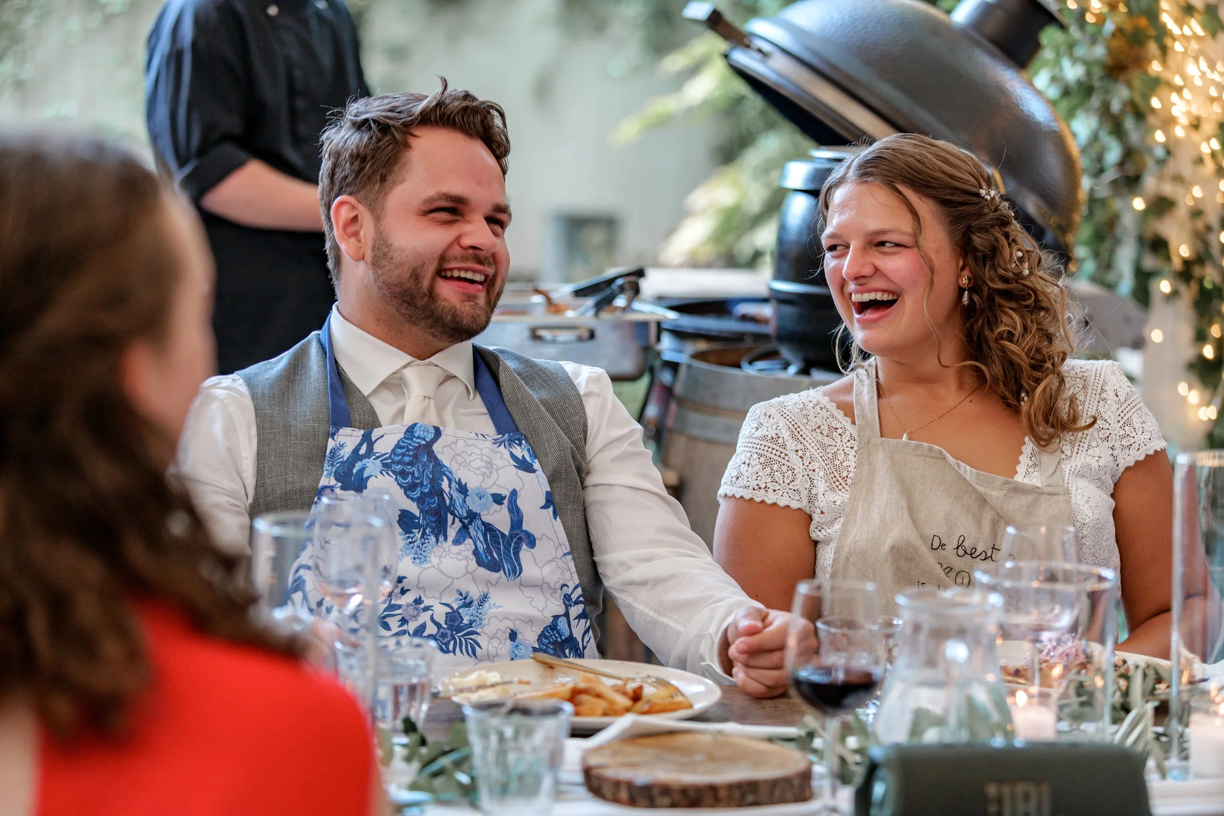 Twee mensen lachen en hebben plezier tijdens een diner op hun trouwdag. Ze zitten aan een tafel met wijn, eten en decoraties, op een feestelijke gelegenheid. Gefotografeerd door professionele fotograaf uit Nederland.