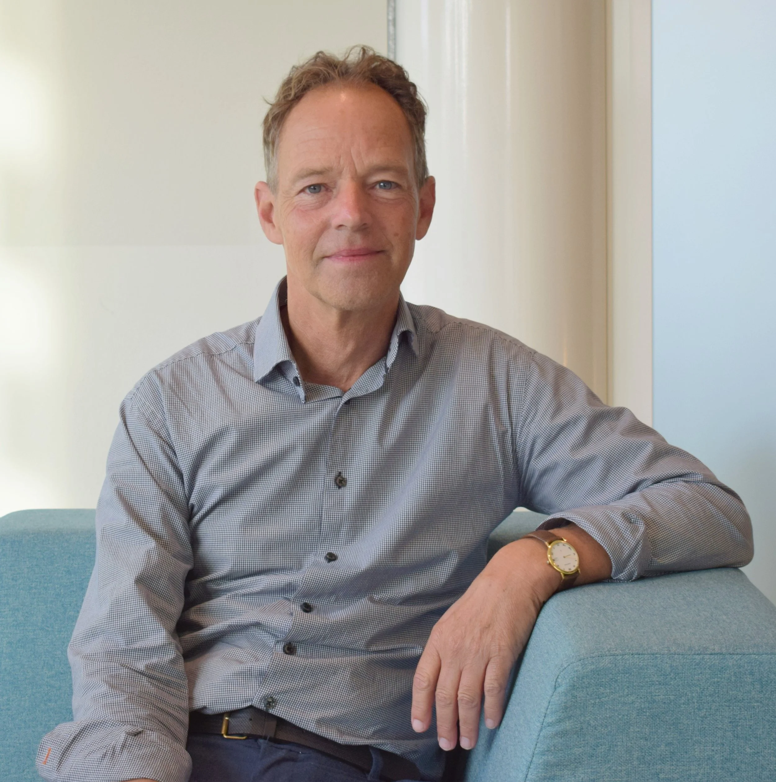 Een man zit op een blauwe stoel, draagt een lichtblauw overhemd en een gouden horloge, en kijkt naar de camera in een kantoorachtige omgeving.