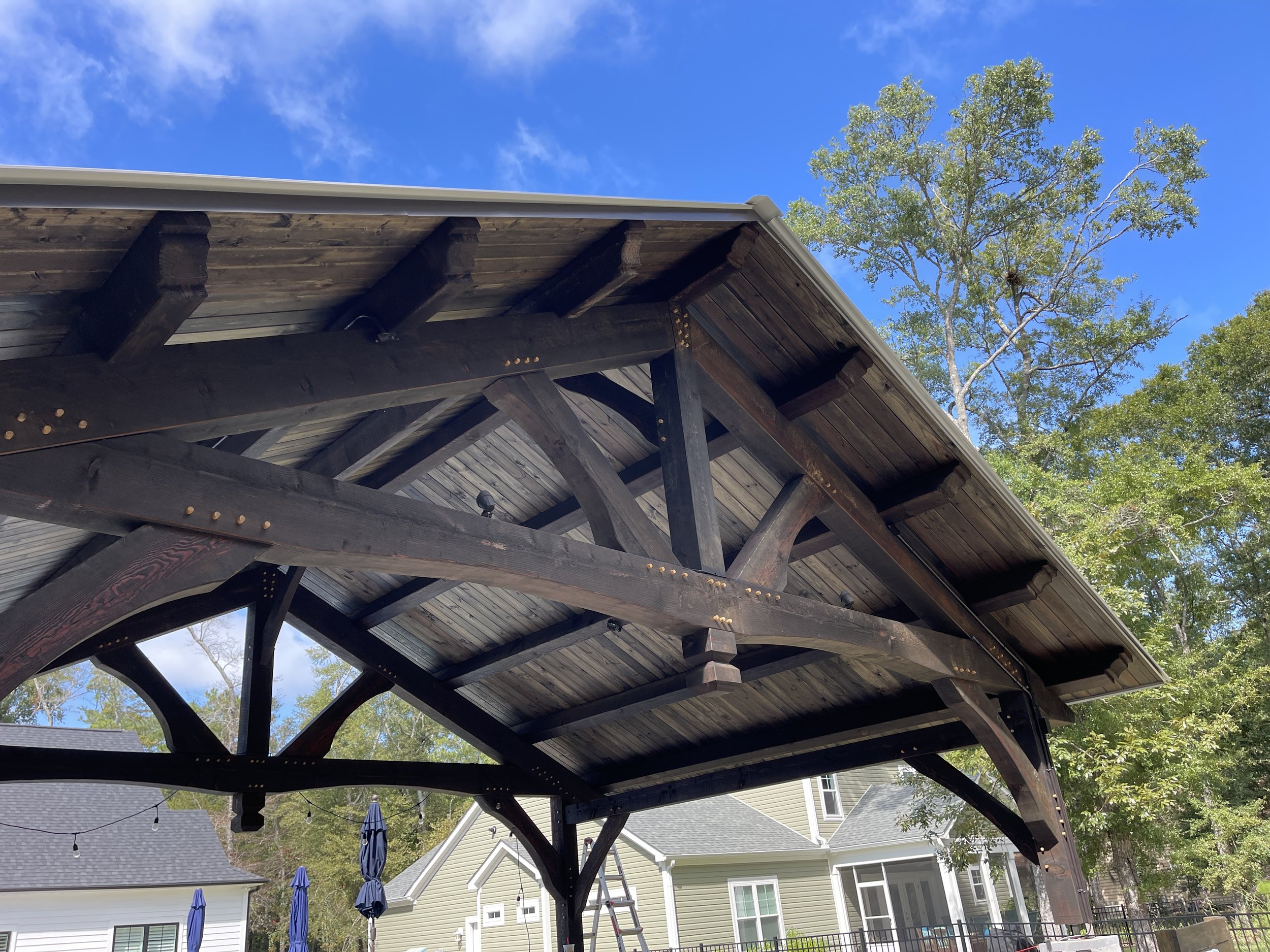 Wooden Timber Frame outdoor patio cover with trees and houses in background under blue sky.