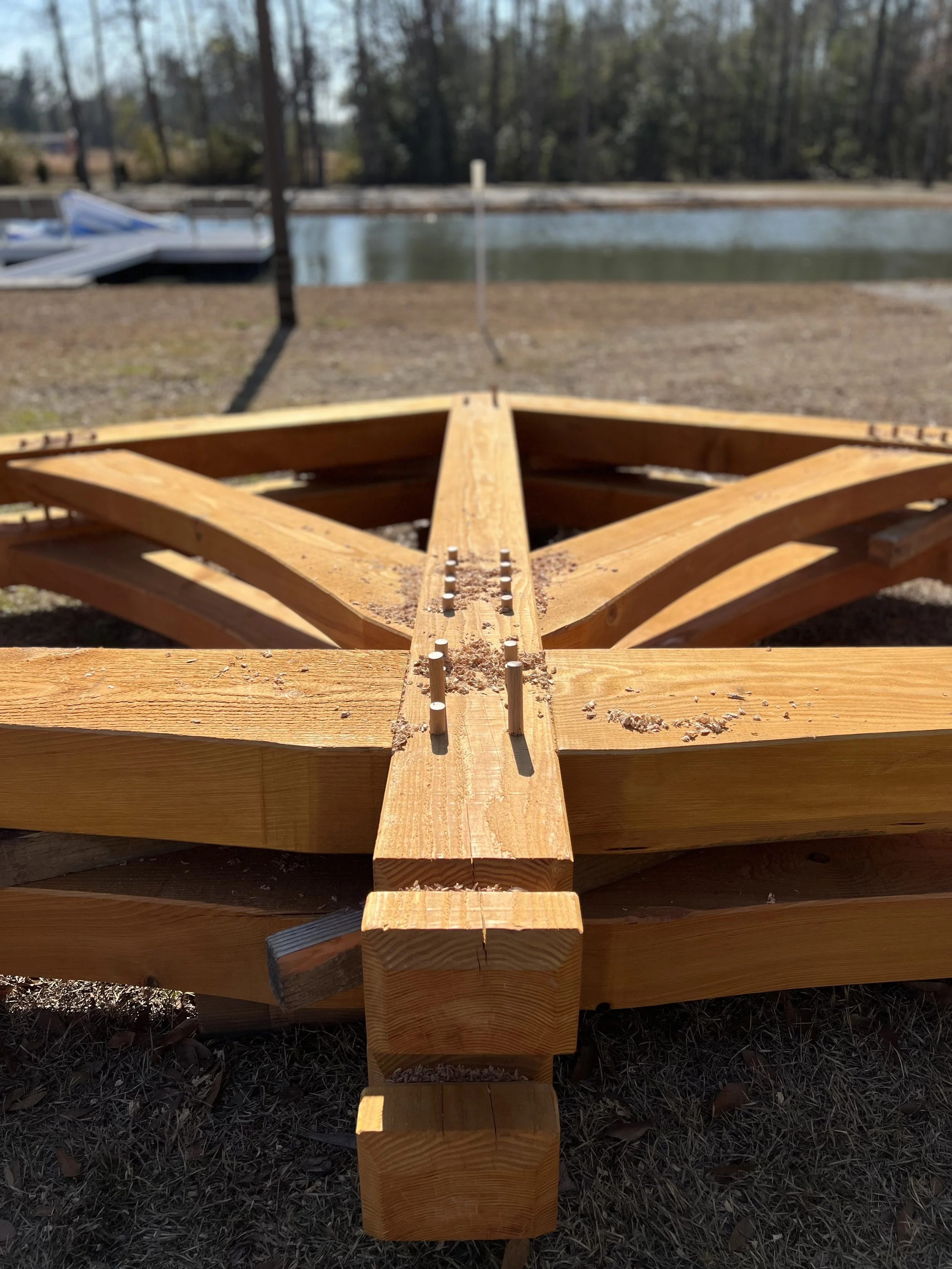 Close-up of a partially assembled wooden timber frame outdoors with a body of water and trees in the background.