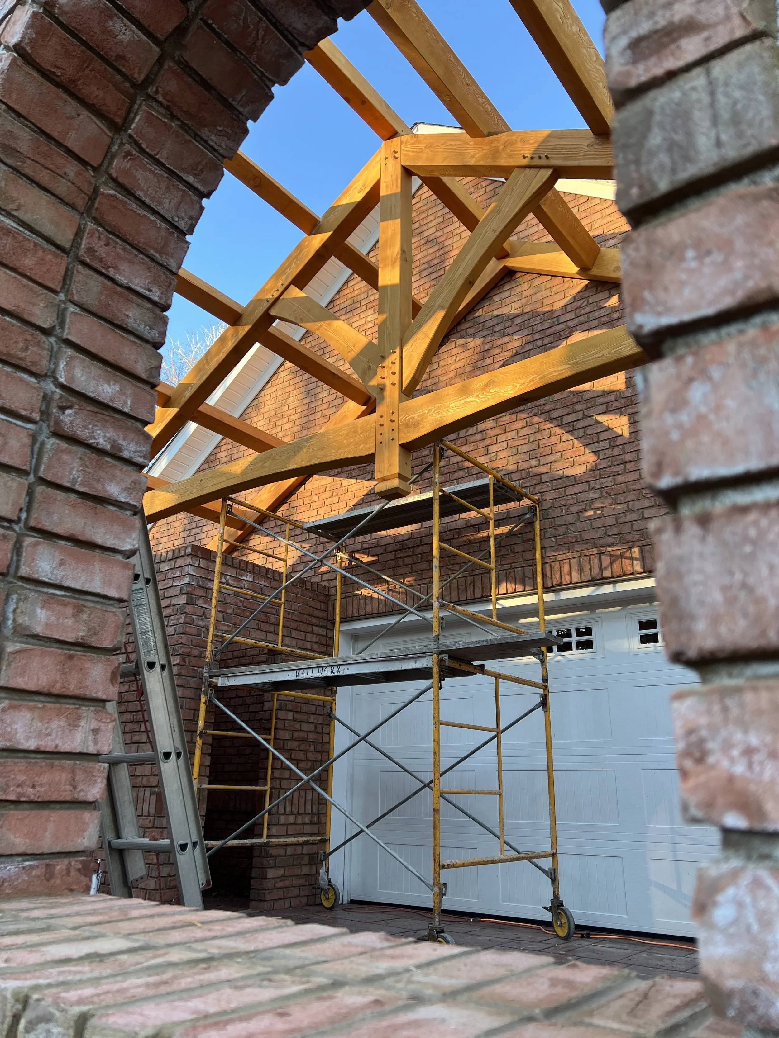 Scaffolding set up in front of a brick house, with wooden roof trusses being installed, viewed from a low angle through an archway.
