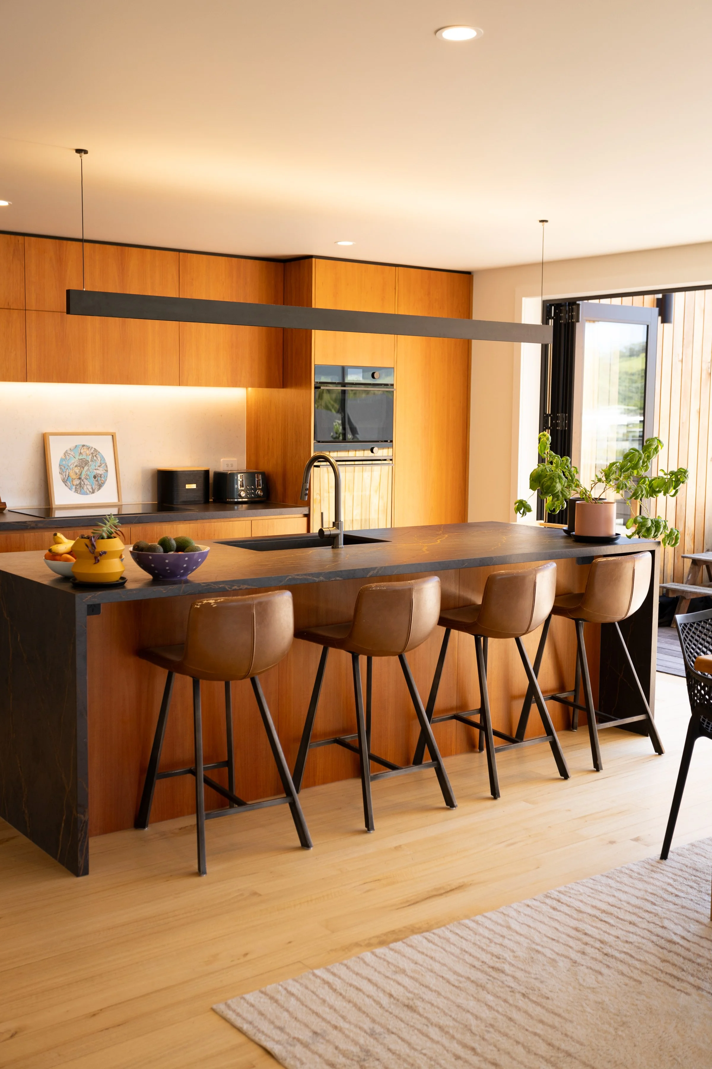 Modern kitchen with wooden cabinetry, a black marble kitchen island with four brown leather bar stools, potted plants, and an open door leading to an outdoor space.