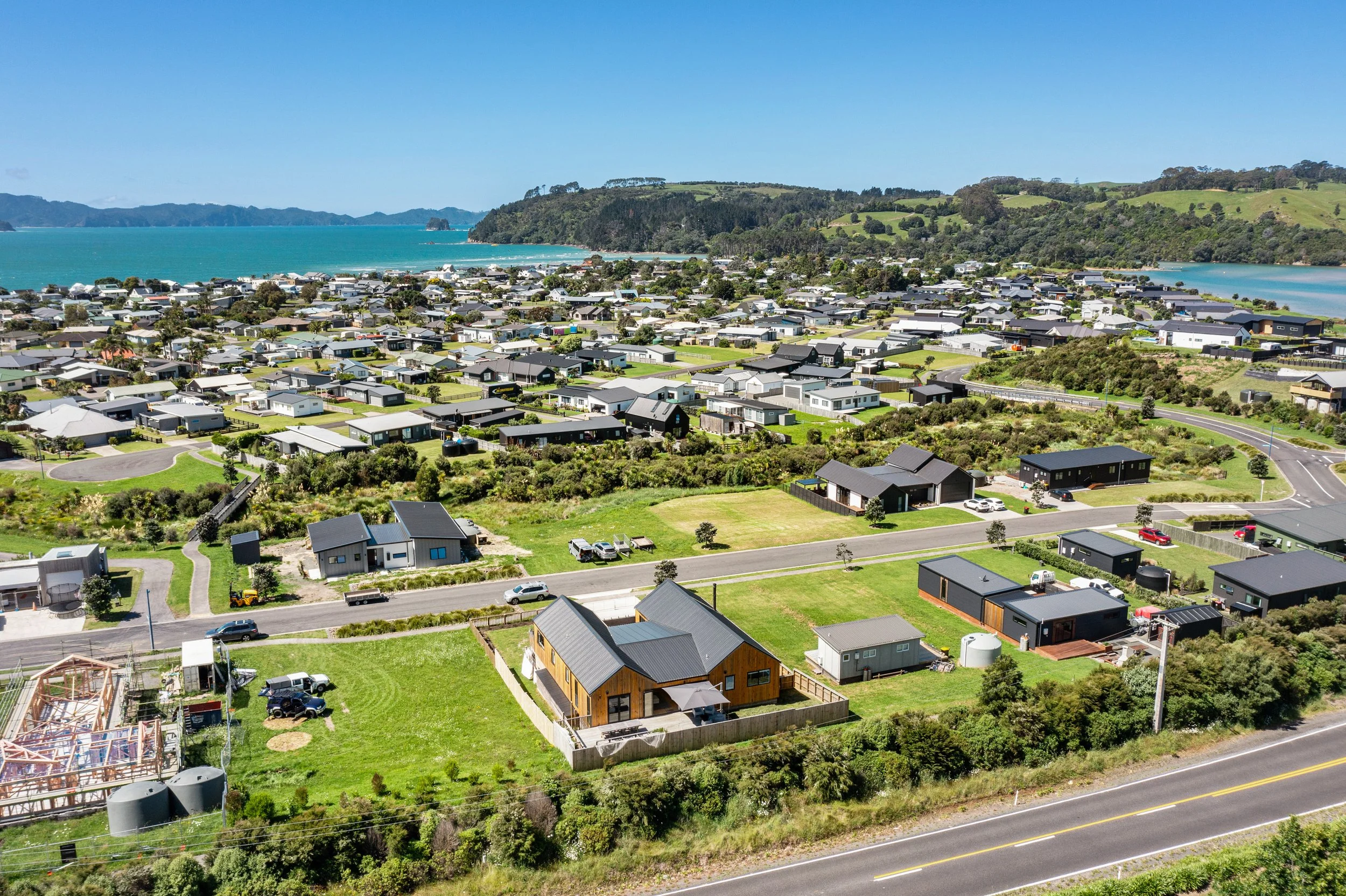 Aerial view of a coastal residential neighborhood with houses, greenery, roads, and a body of water in the background.