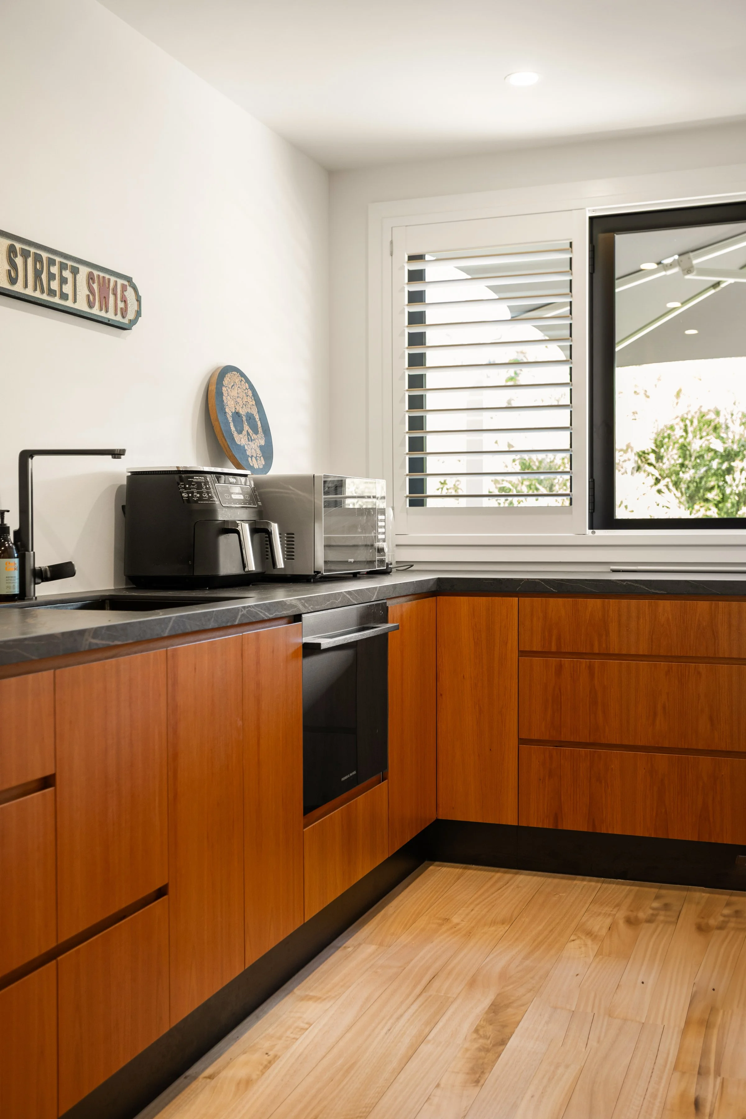 Kitchen with wooden cabinets, black countertop, small black stove, window with white shutters, and outdoor view with trees.
