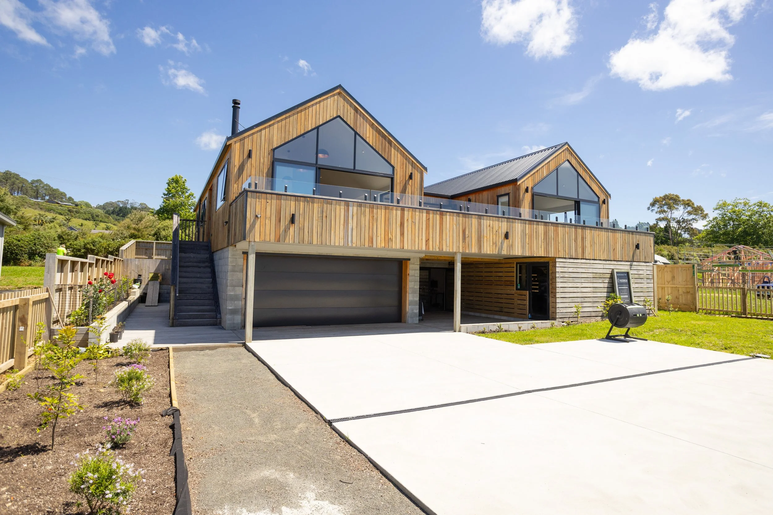 Modern wooden house with two levels, large glass windows, and a spacious driveway under a blue sky with clouds.