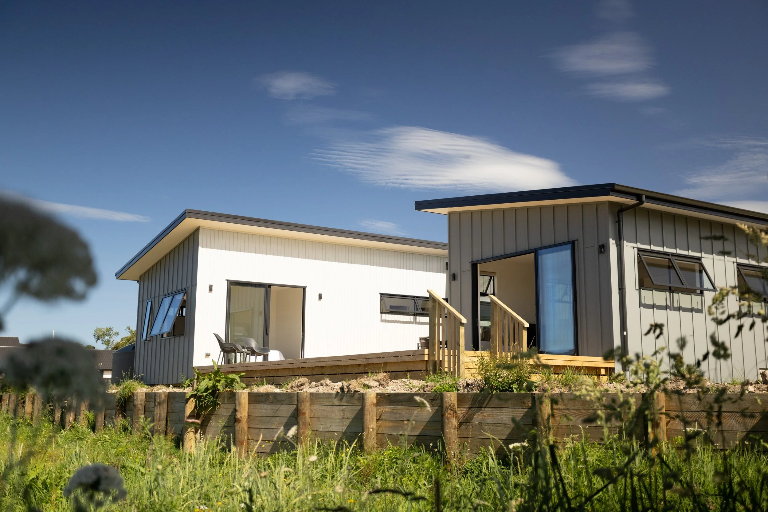 Modern white houses with large windows and small balconies on a grassy hillside under a blue sky with clouds.