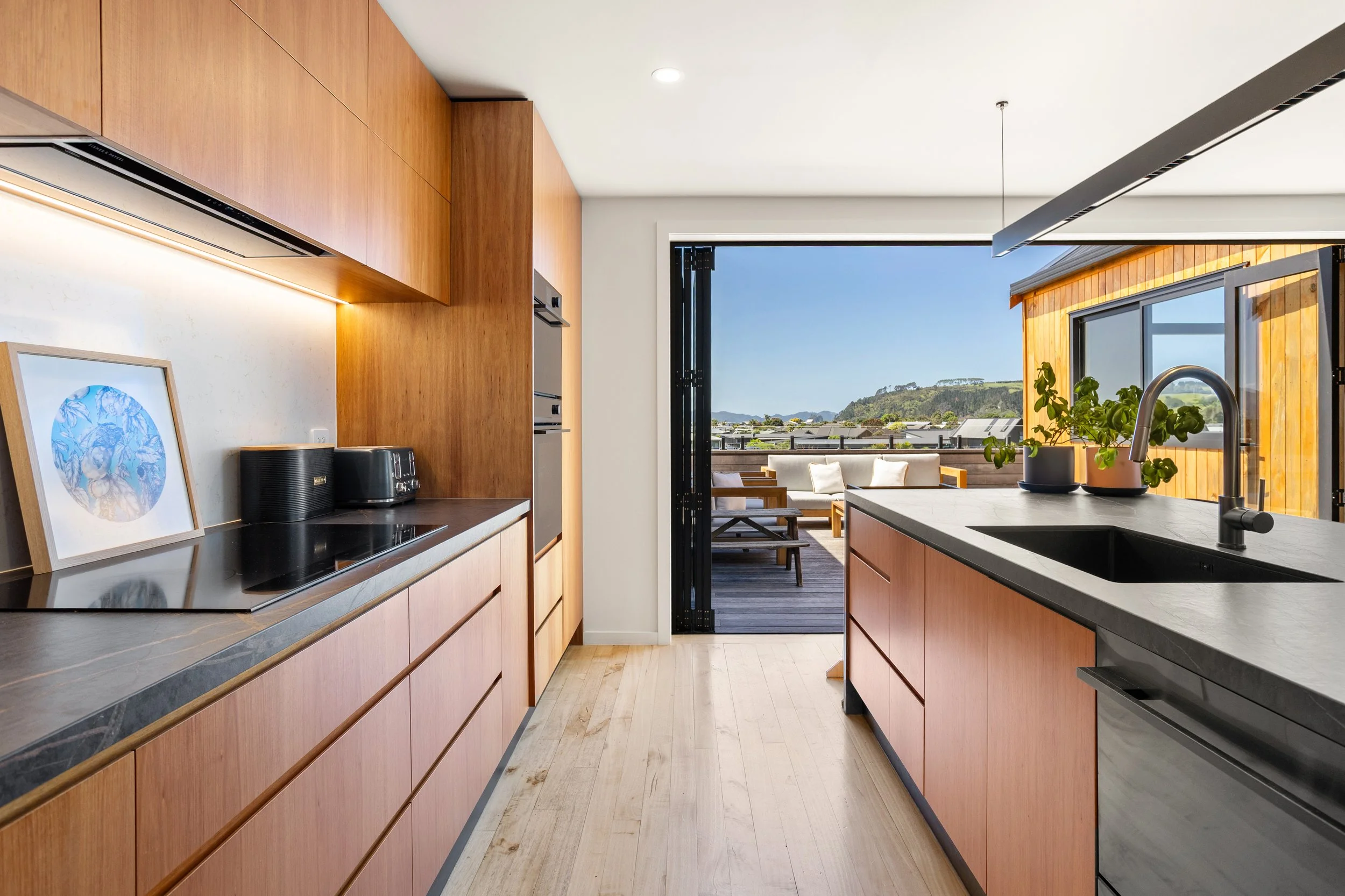 Modern kitchen with wooden cabinets, black countertops, and a view of an outdoor patio with seating and greenery.