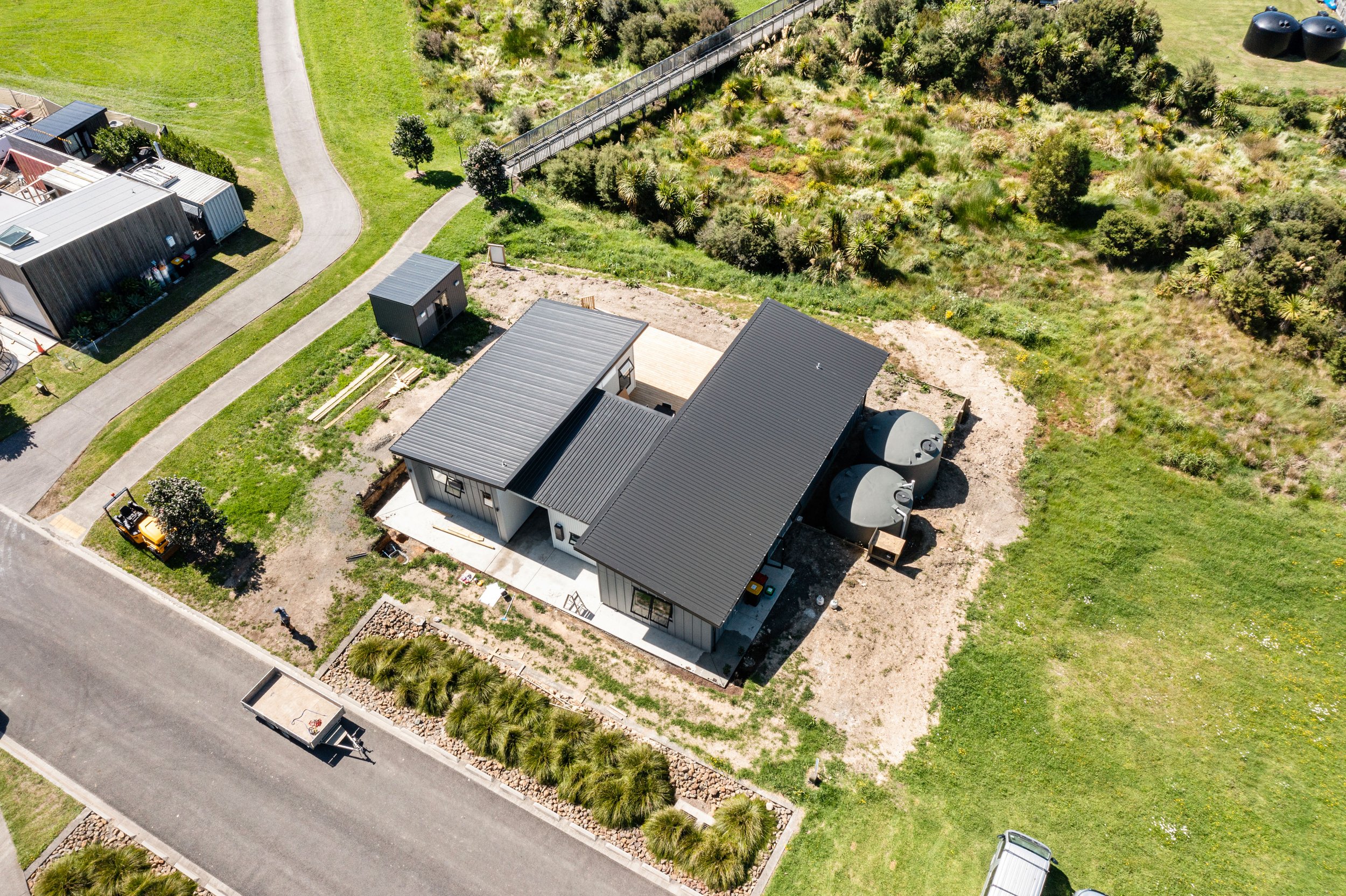 Aerial view of a modern house with a metal roof, surrounded by a green lawn, trees, and a neighborhood street with other houses and a trailer nearby.
