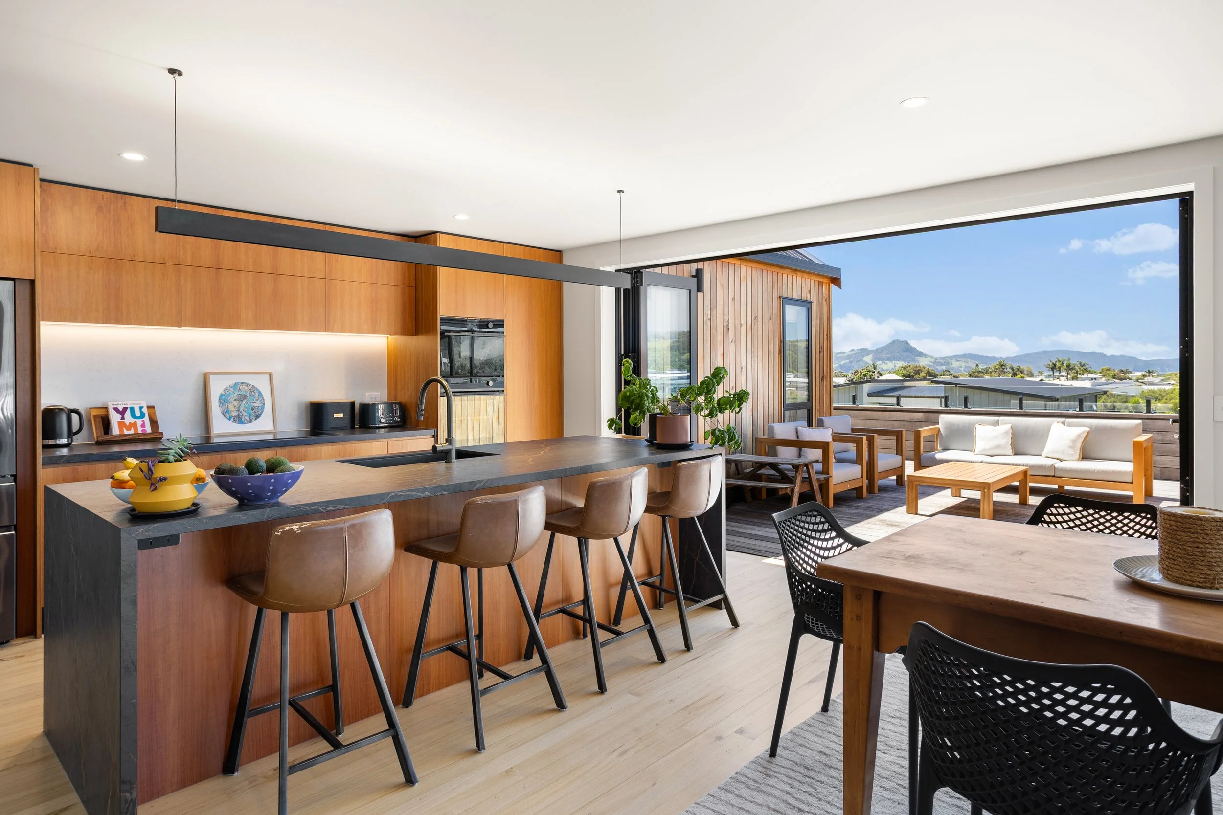 Open-concept living space with a kitchen island, bar stools, and a sitting area on a balcony with outdoor furniture and mountain view.
