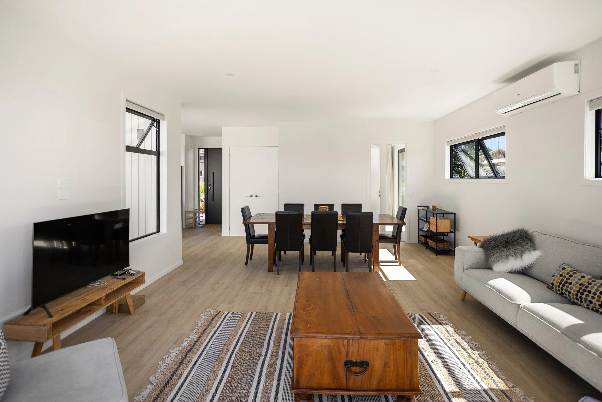 Living room with white walls, hardwood floor, a gray sofa with a fuzzy pillow, a wooden coffee table, a patterned rug, a flat-screen TV on a wooden stand, and a dining table with black chairs. Multiple windows allow natural light into the space.