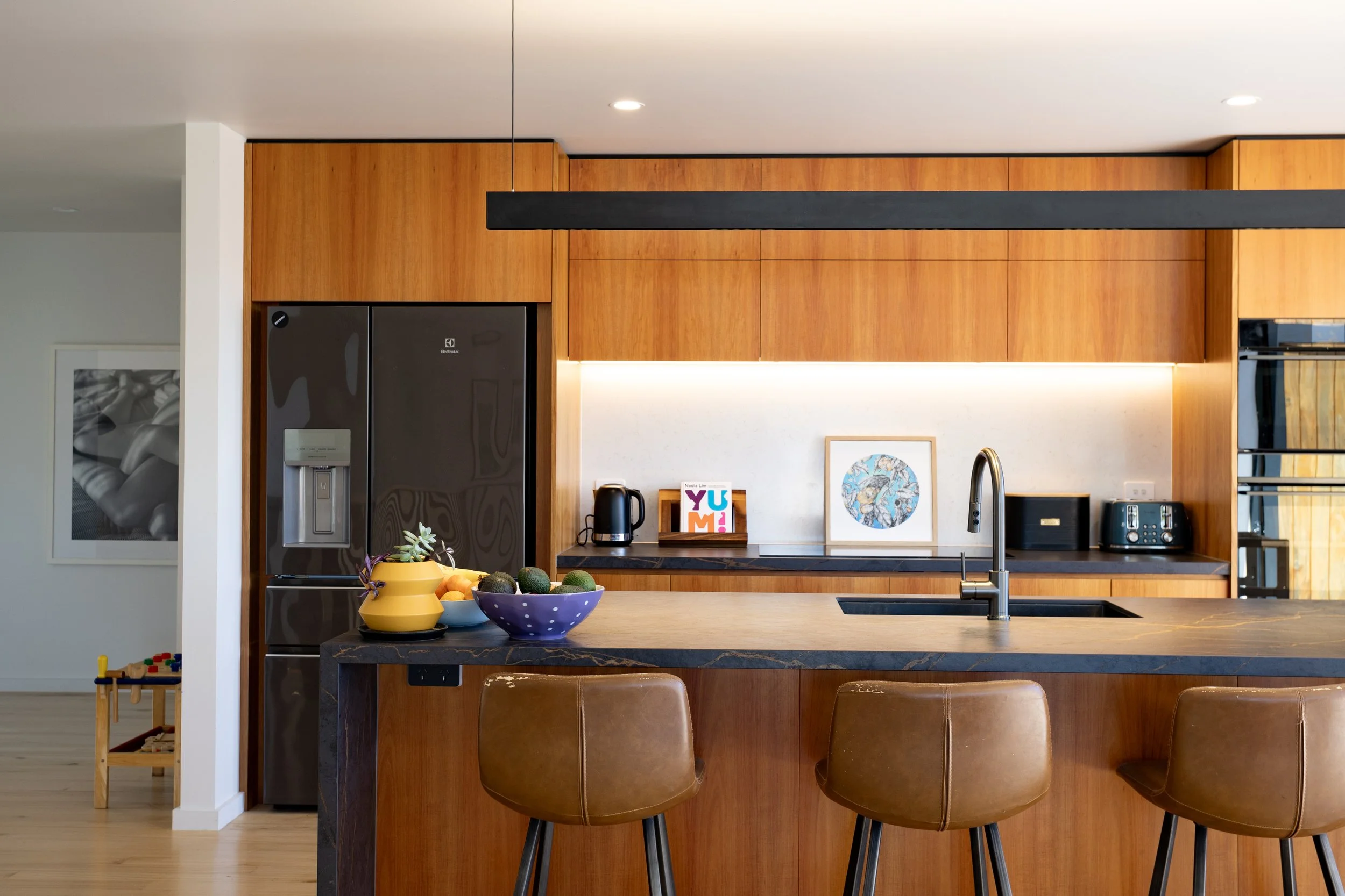 Modern kitchen with wooden cabinetry, black countertop, and brown bar stools. Contains fruit bowls, a coffee maker, toaster, and framed art. Refrigerator and other appliances in the background.