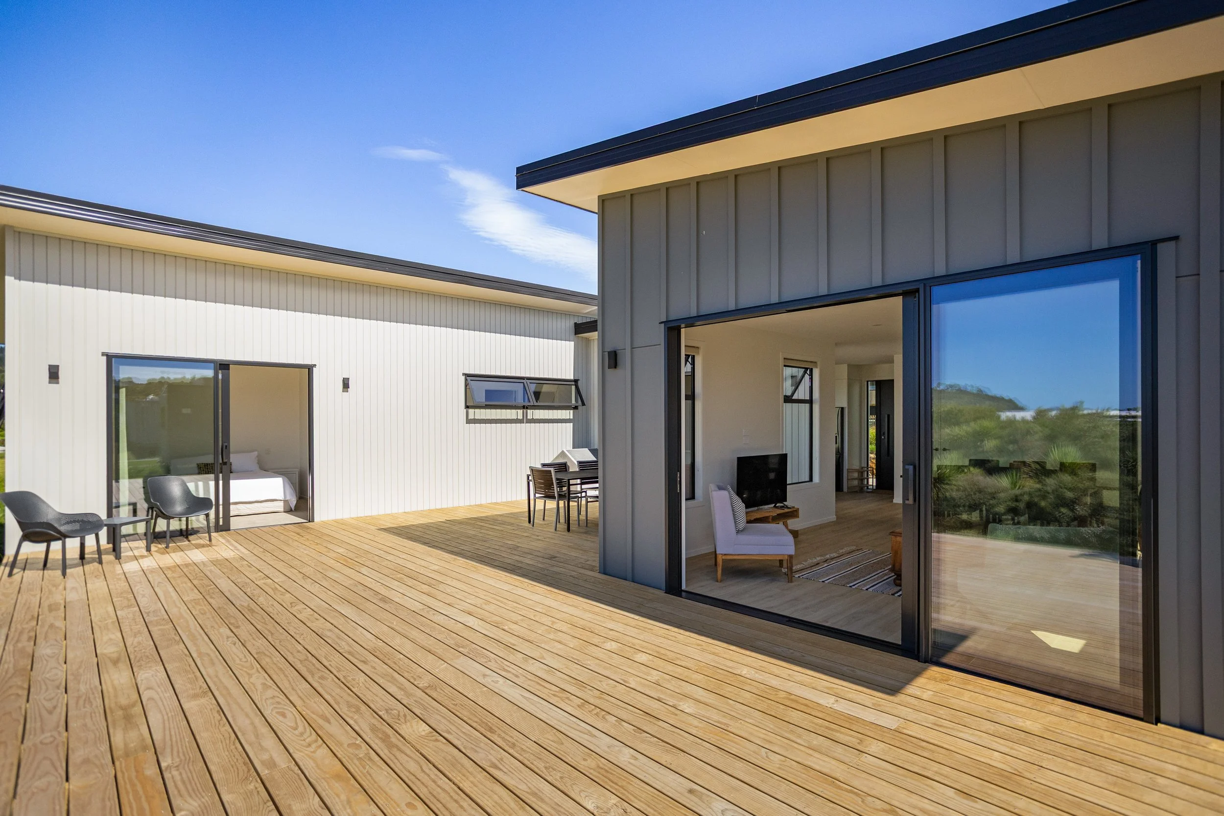 Modern house with a wooden deck, large sliding glass door, and a living room visible inside. The house has a gray exterior and is set against a clear blue sky.