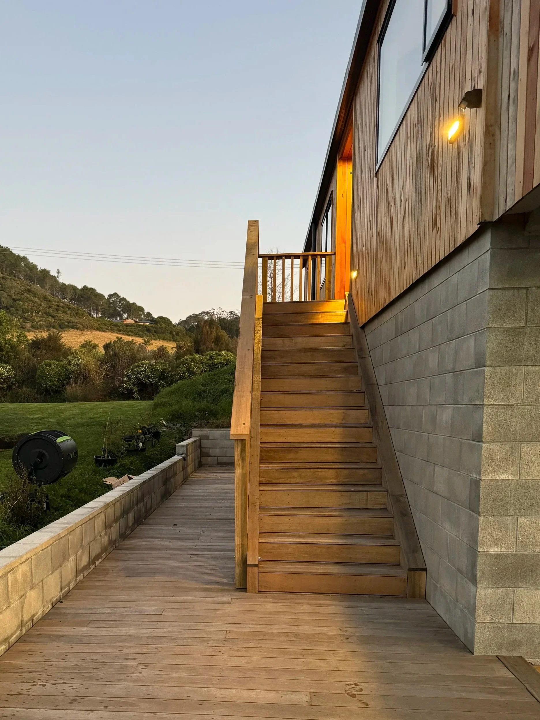 Wooden staircase and deck outside a modern house with large windows and exterior lighting, overlooking a green landscape during sunset.