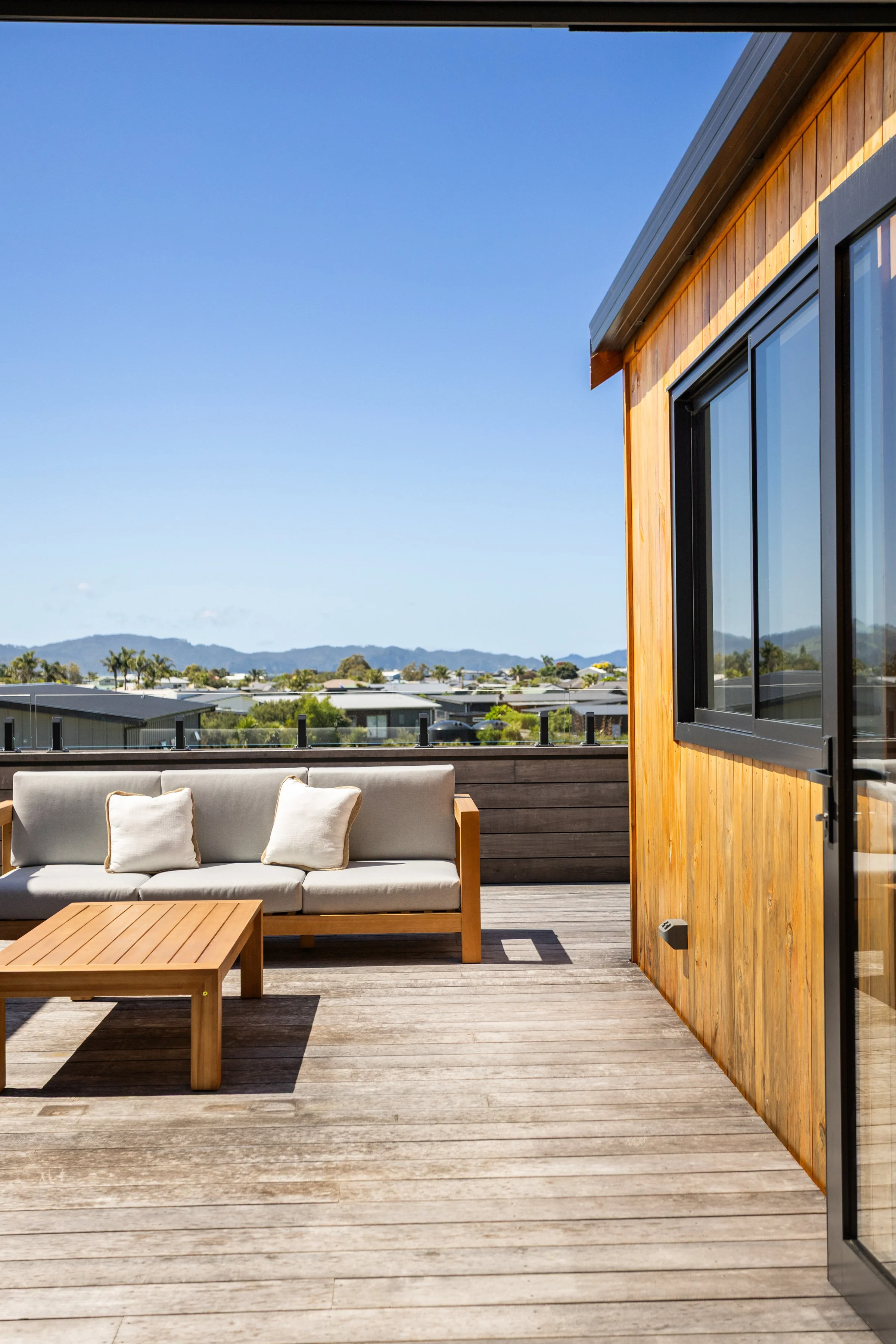 Outdoor balcony with a wooden floor and wall, beige sofa with pillows, a wooden coffee table, and a view of rooftops and mountains under a clear blue sky.