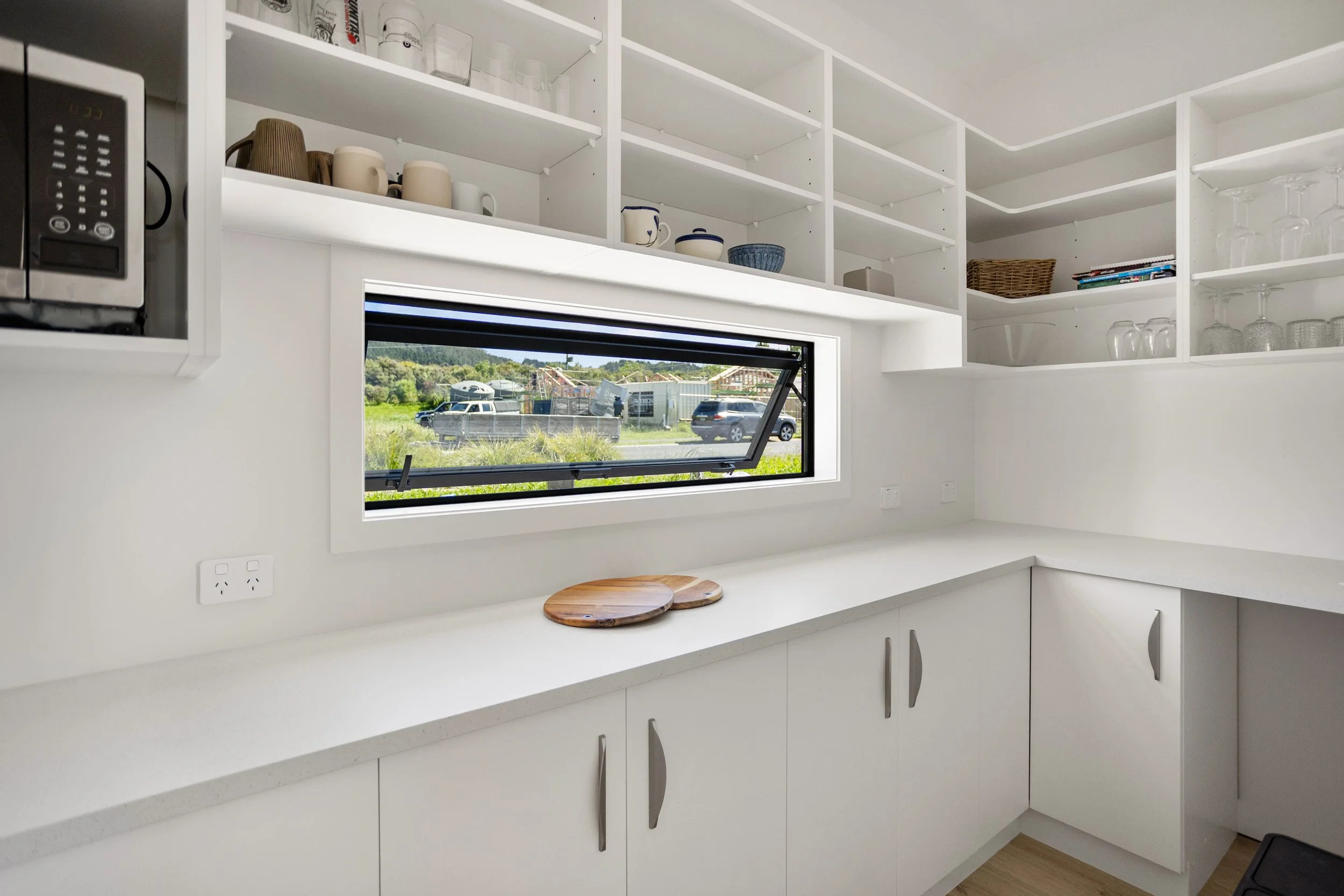 Modern kitchen with white cabinets, open shelves with dishes and glassware, a window showing outdoor scenery, and a countertop with two wooden cutting boards.