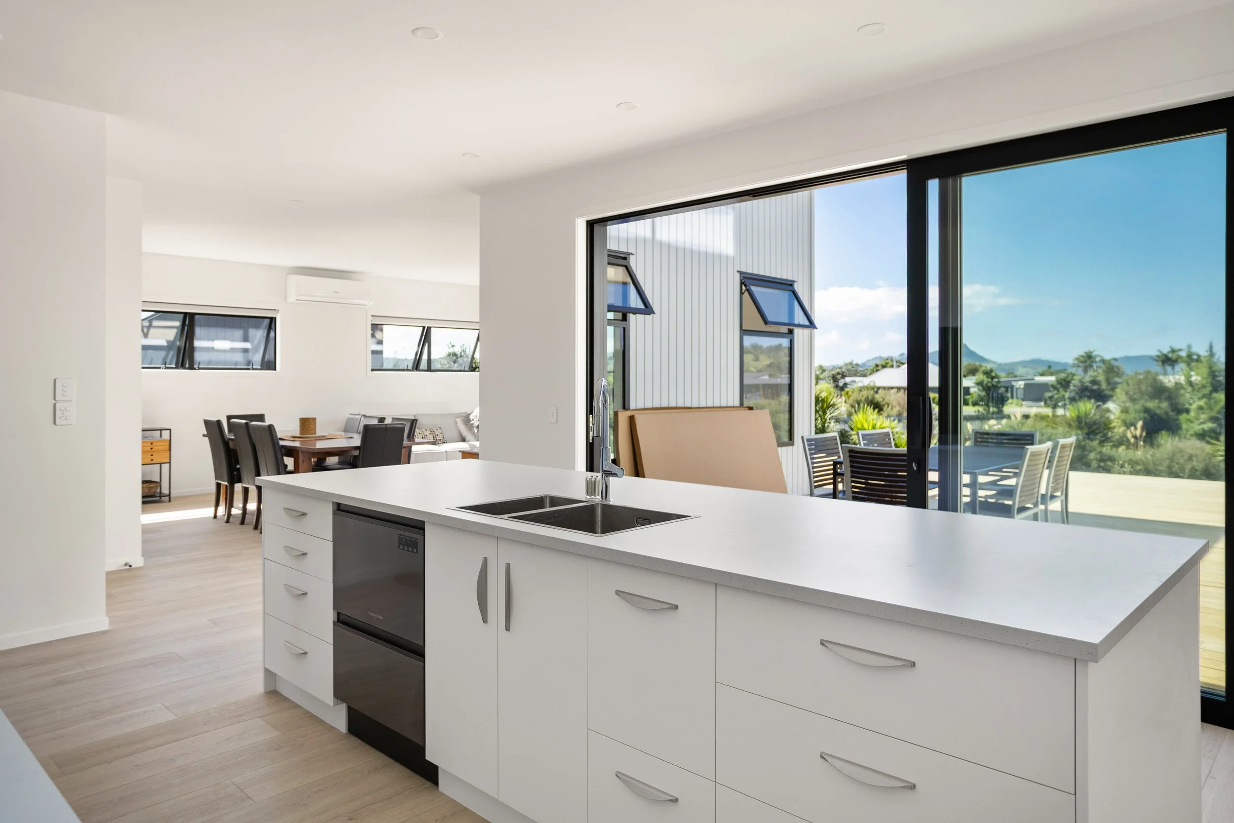 Modern open-concept kitchen with white cabinetry, a large island with a sink, and a view of an outdoor patio and lush landscape through sliding glass doors.