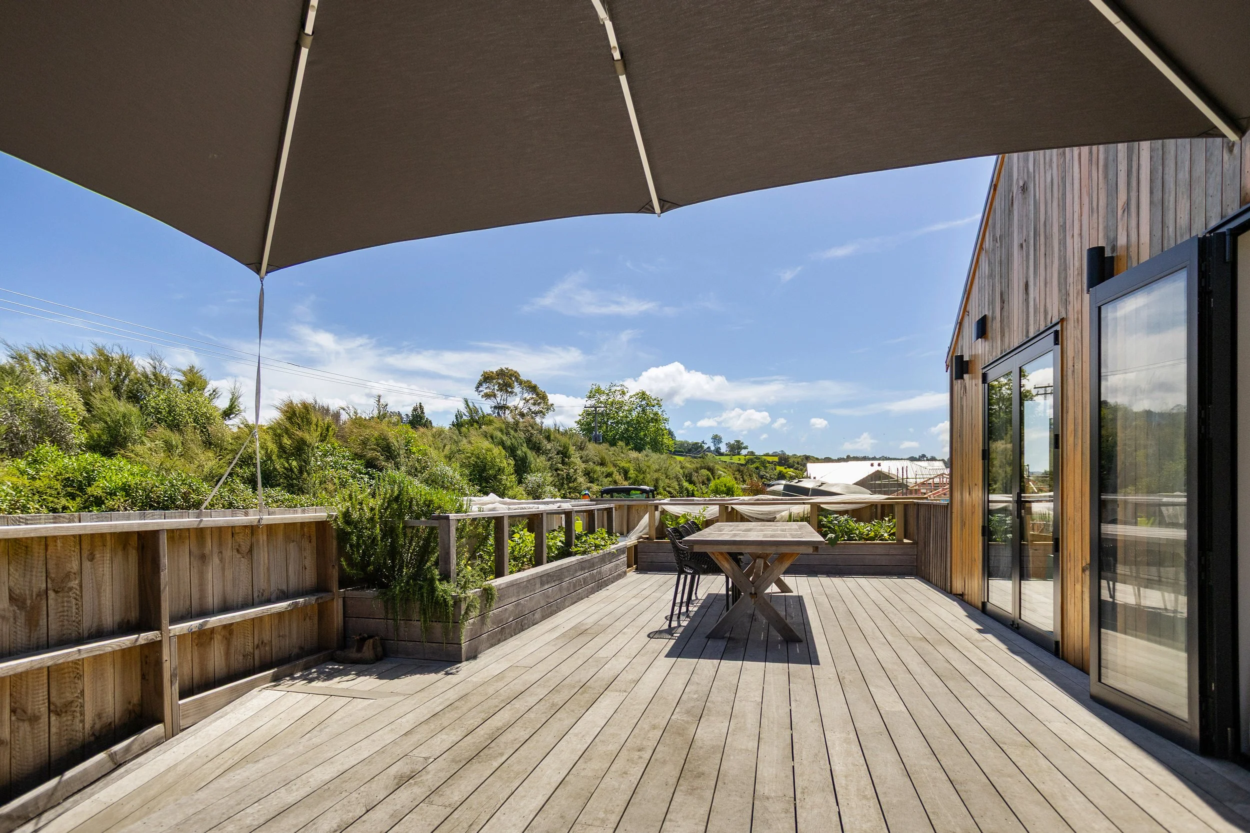 Outdoor wooden deck with a large brown umbrella, outdoor table, and chairs, overlooking lush greenery and a blue sky with scattered white clouds.