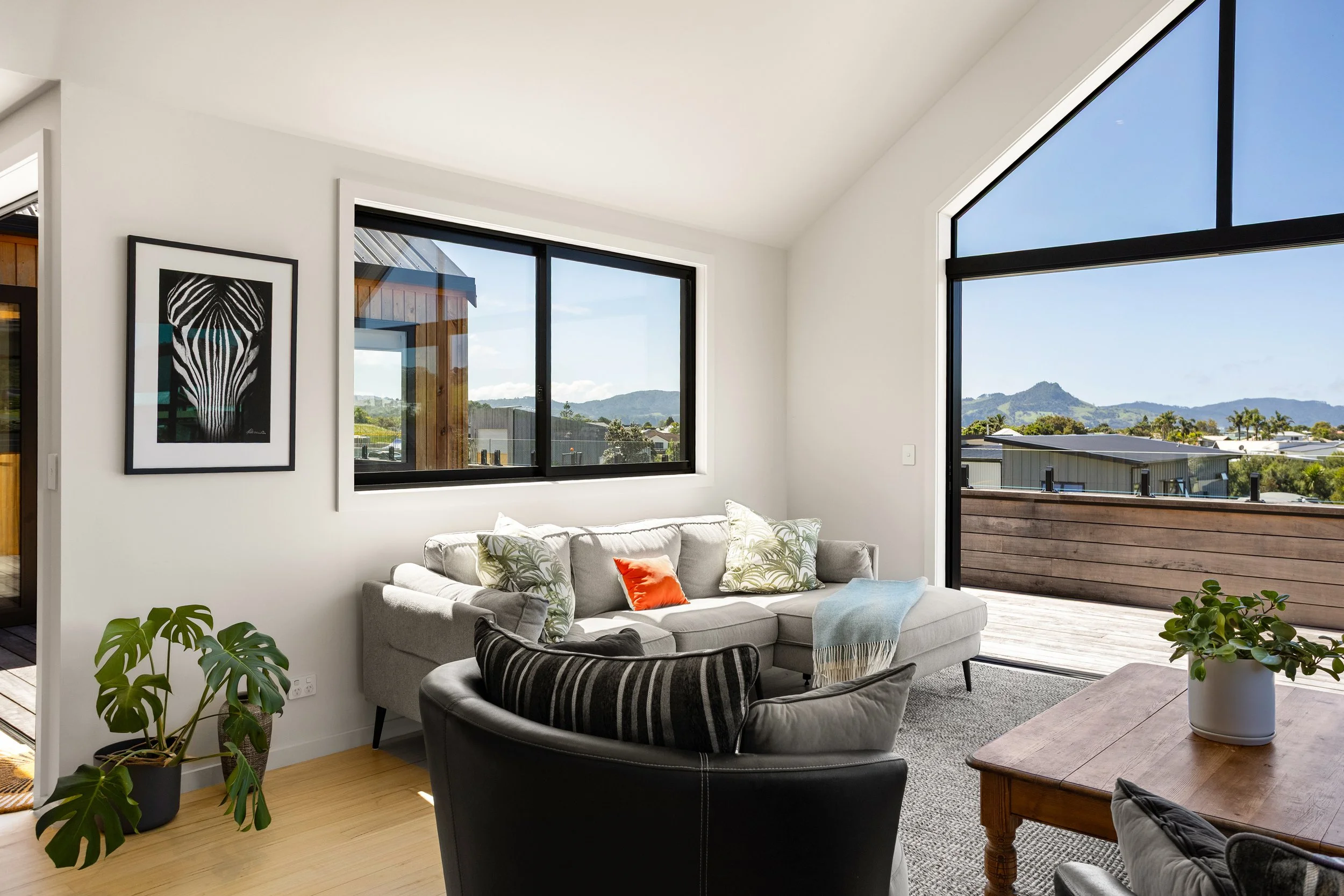 Living room with large windows, gray sofa with pillows, wooden coffee table with a potted plant, and a balcony with a mountain view