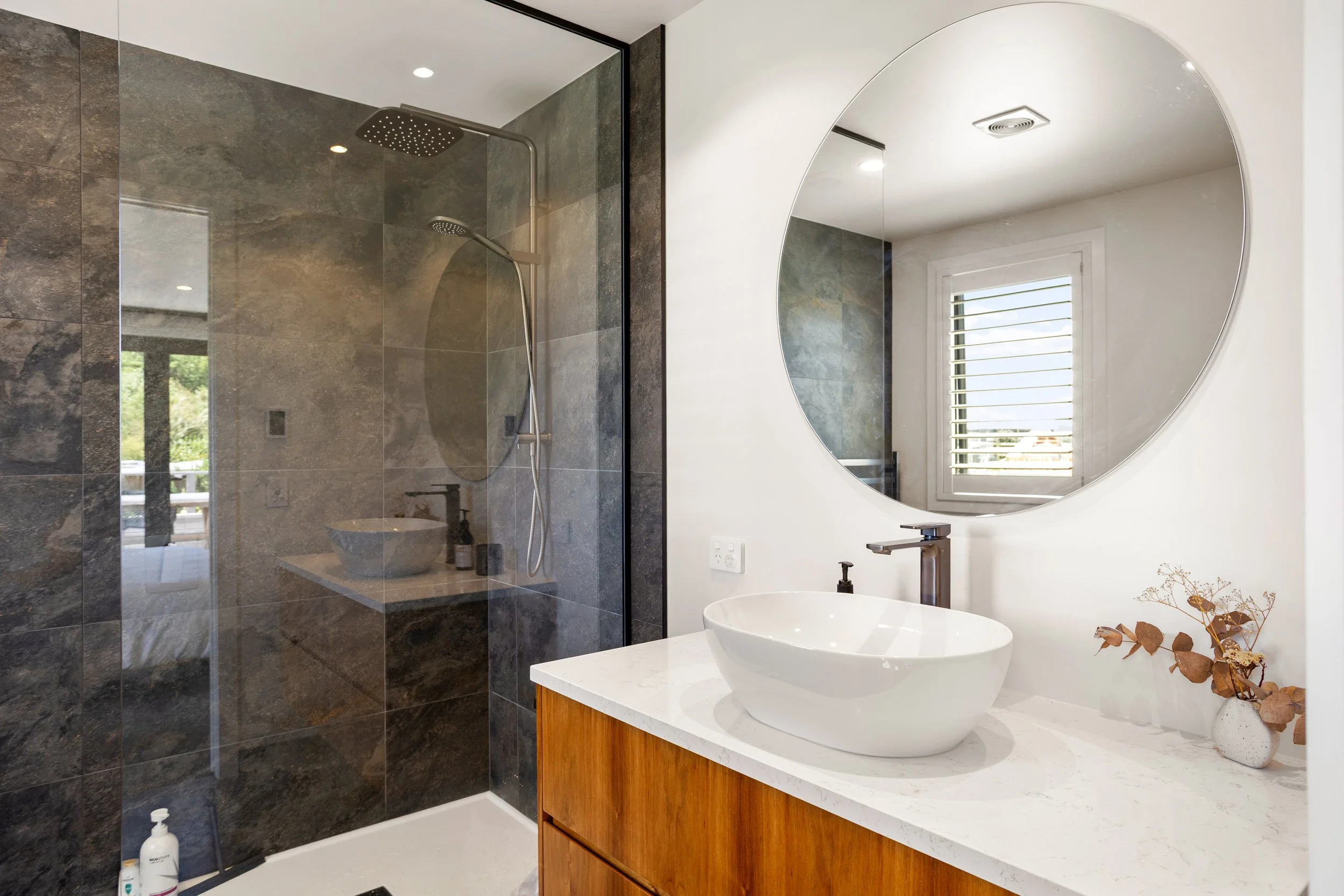 Modern bathroom with a walk-in shower with dark tiles, a white countertop with a vessel sink, round mirror, and a window with white blinds.
