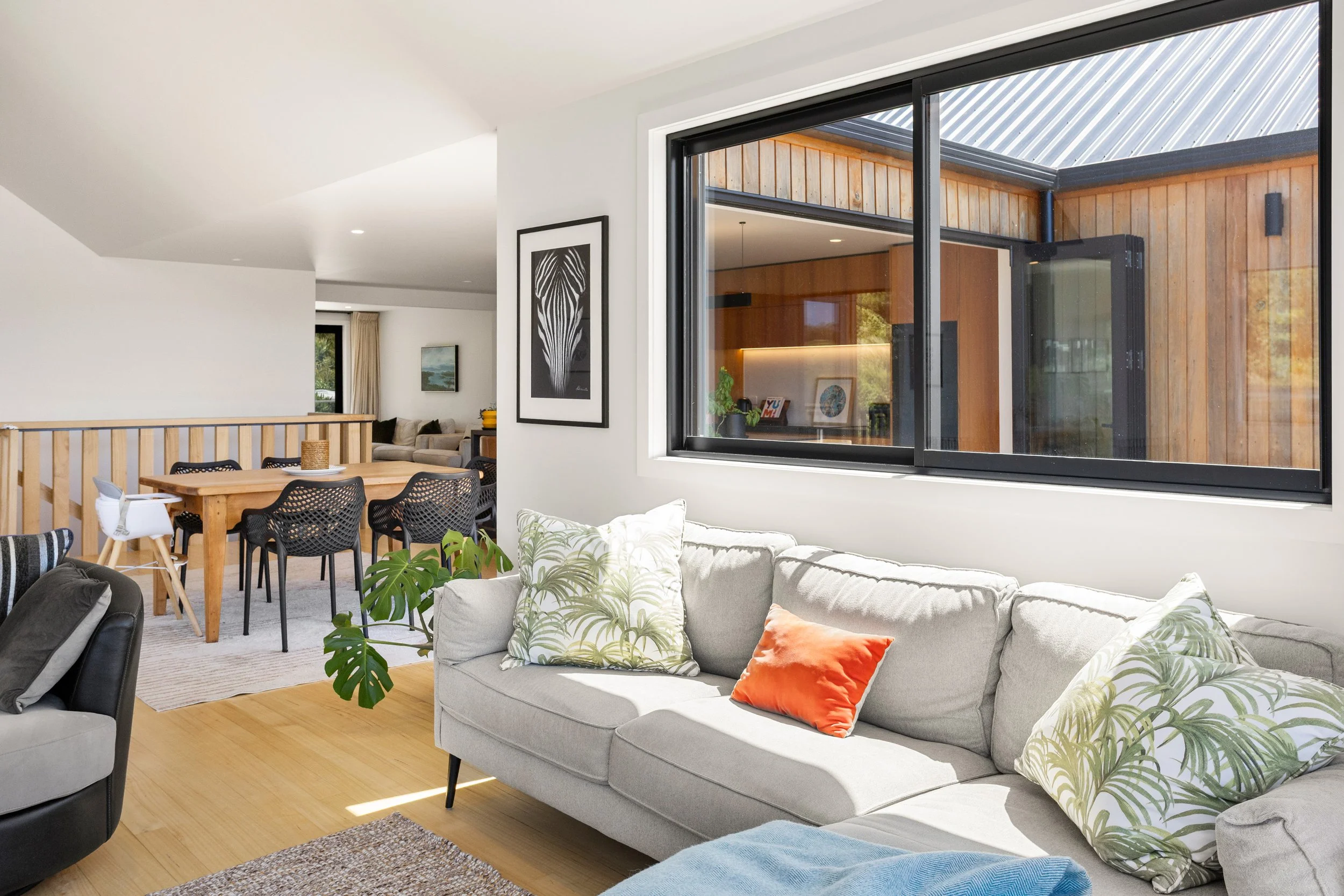Living room with a beige sofa decorated with green and white patterned pillows and an orange pillow, a plant, and a large window showing an outdoor patio with wooden walls.