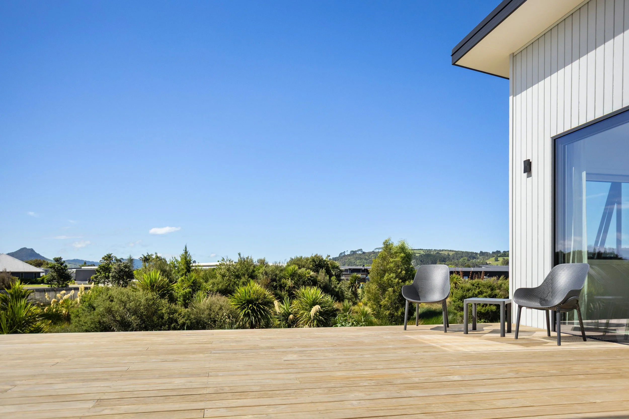 Modern outdoor patio with two gray chairs and a small table, overlooking lush greenery and distant hills under a clear blue sky.