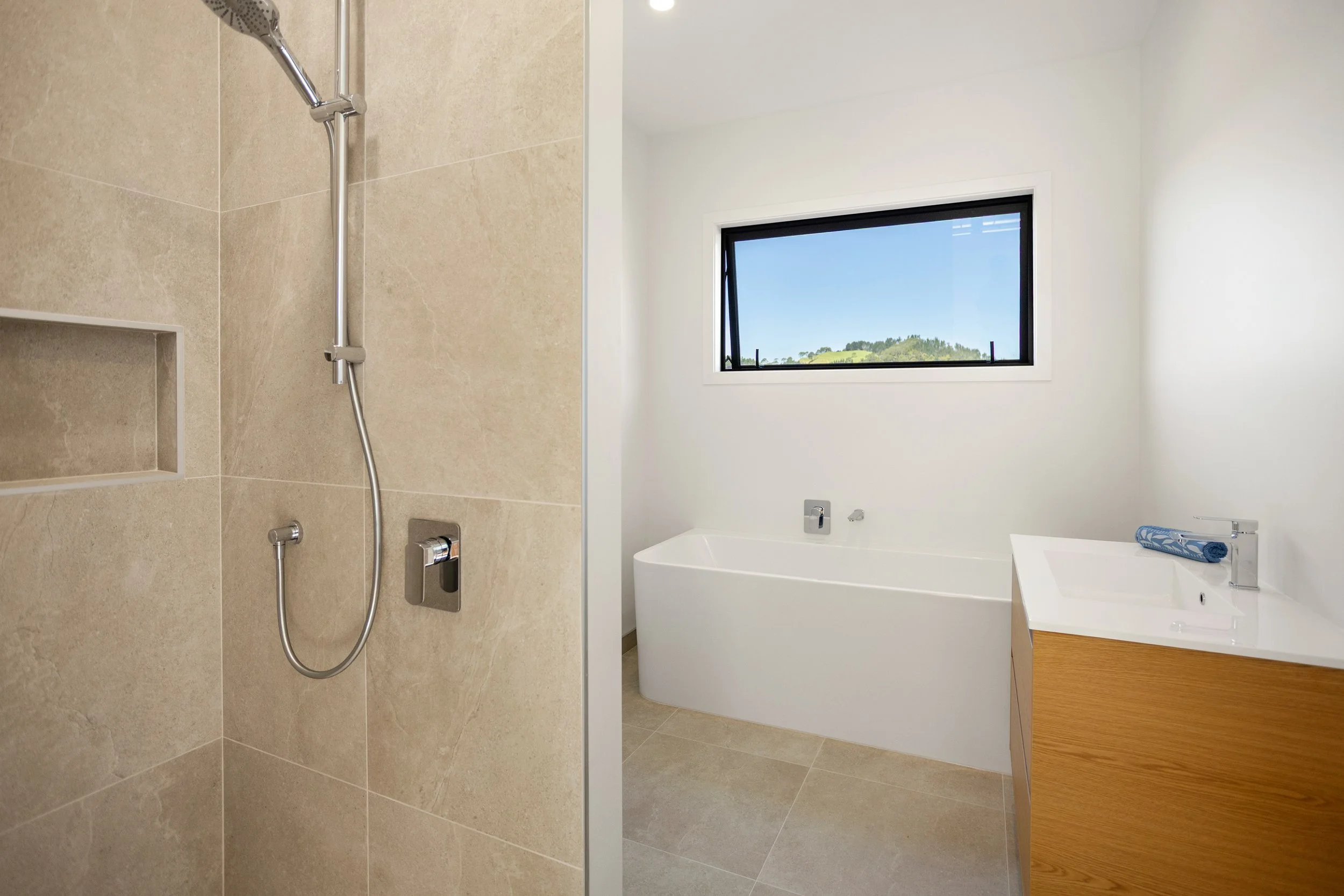 Modern bathroom with a beige tiled shower area, a bathtub beneath a window showing a landscape view, and a white vanity with a sink and a rolled-up towel.