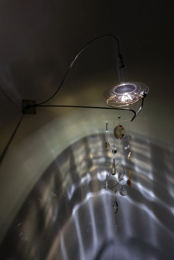 Wall-mounted fixture made of bent steel and brass accents with vintage glassware: a pink depression plate and an assortment of nineteen crystals. LED-lit, it casts white light in a faceted pattern.