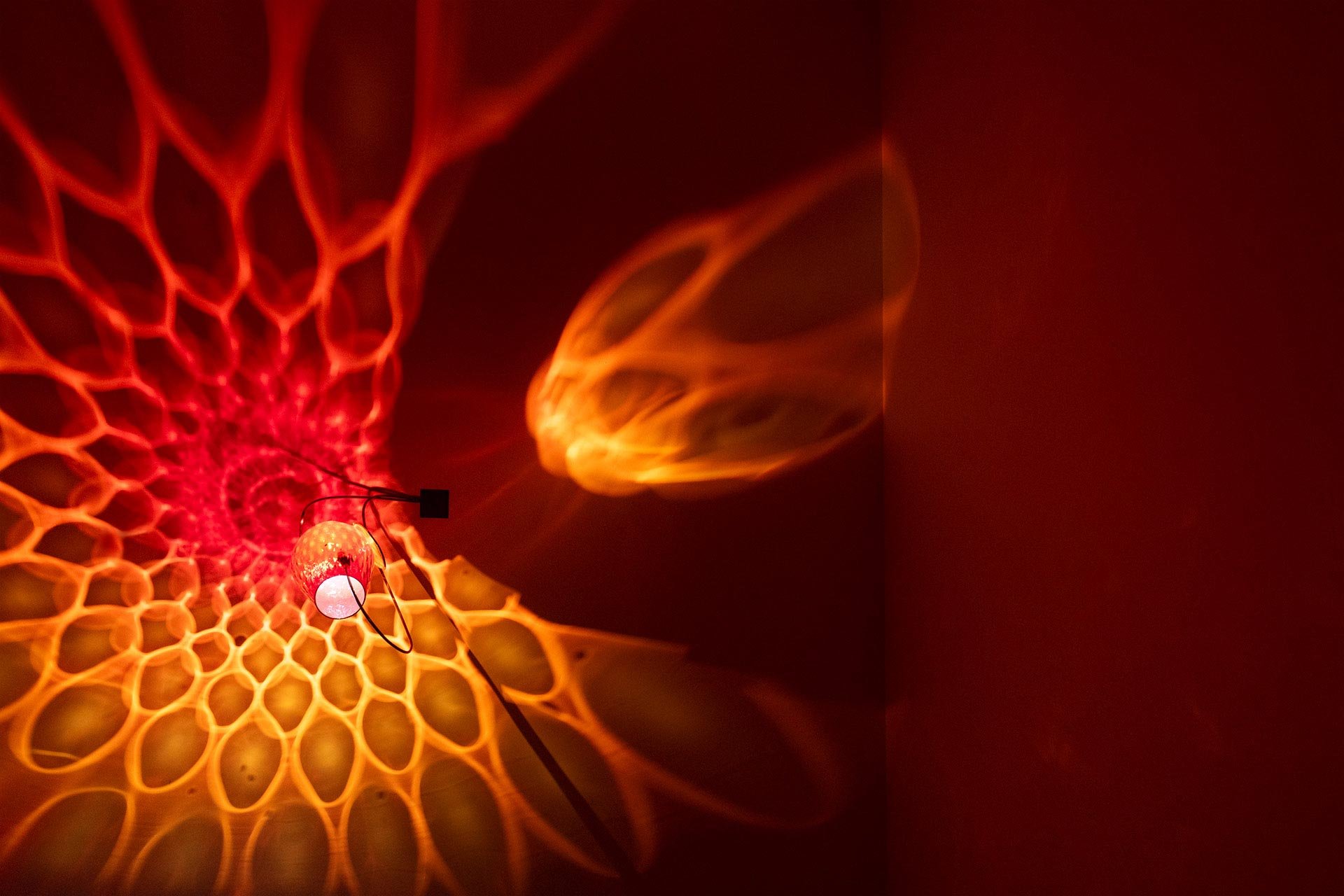 Illuminated, wider view of the fixture displays red and orange light in a mandala-like pattern.