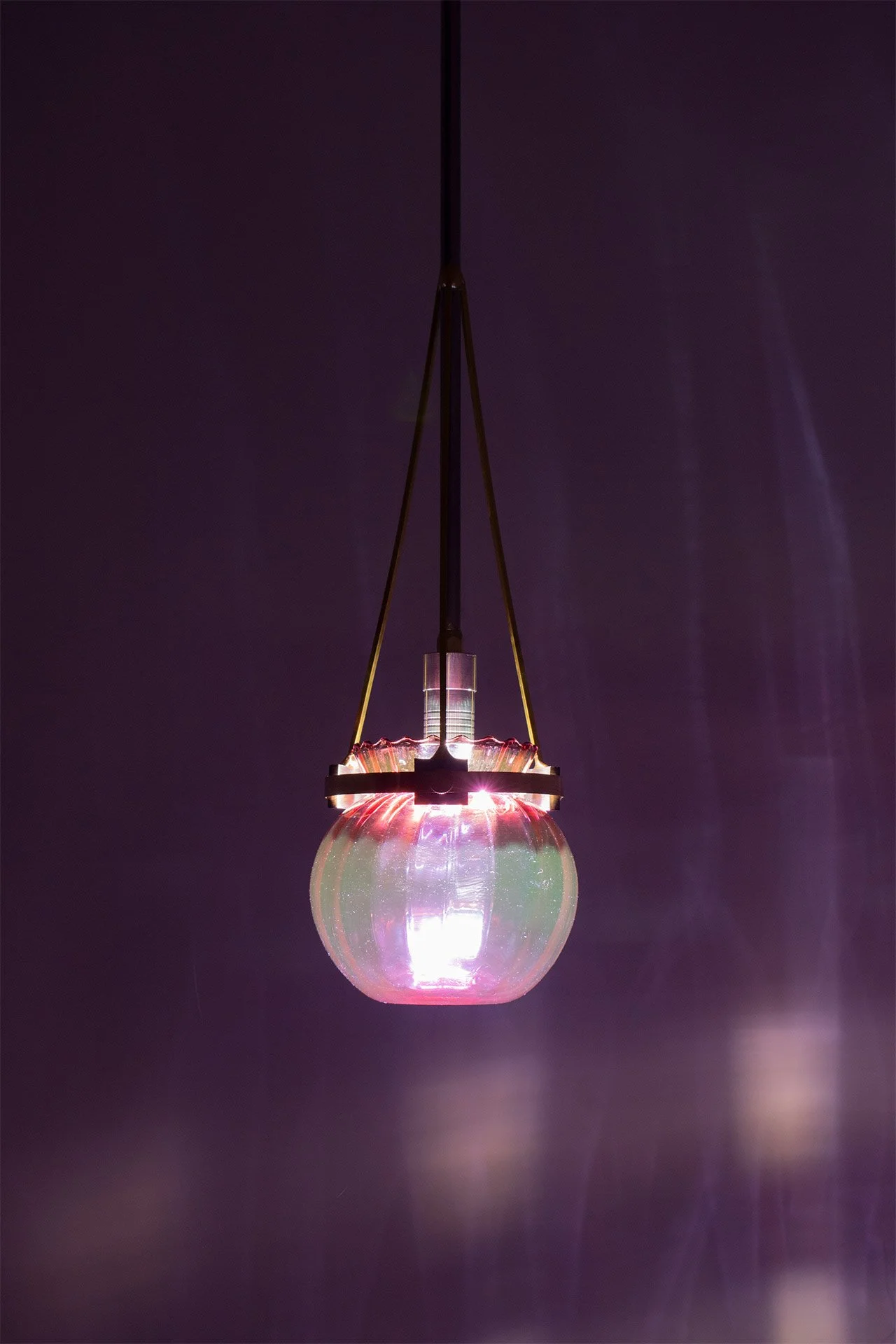 Ceiling pendant fixture made of bent steel piping, brass accents, and a cranberry vintage ribbed glass jar. LED-lit, it casts vertical lines of pink light.