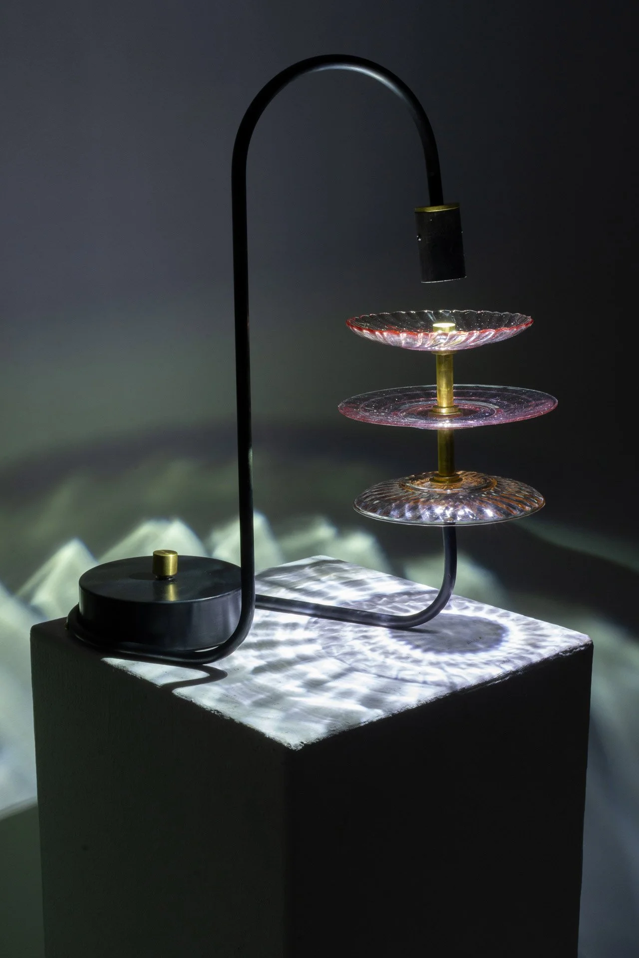 Table lamp made of bent steel piping, a steel circular base, brass accents, and three vintage pink vintage depression glass plates. LED-lit, it casts white light in a ribbed, ethereal pattern.