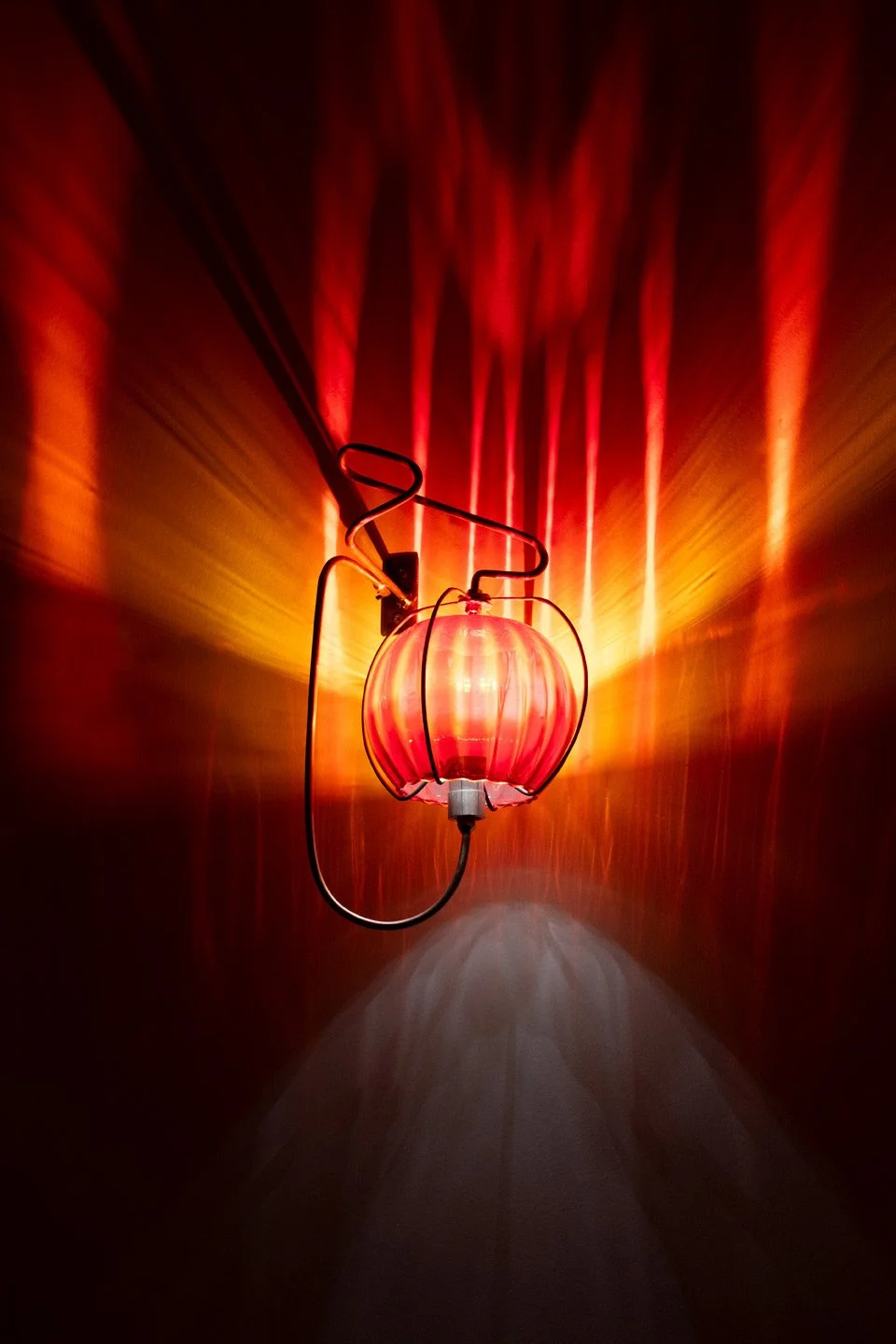 Wall-mounted fixture made of bent steel, brass accents, and a red vintage ribbed glass vase. LED-lit, it casts red and orange light that splits linearly.