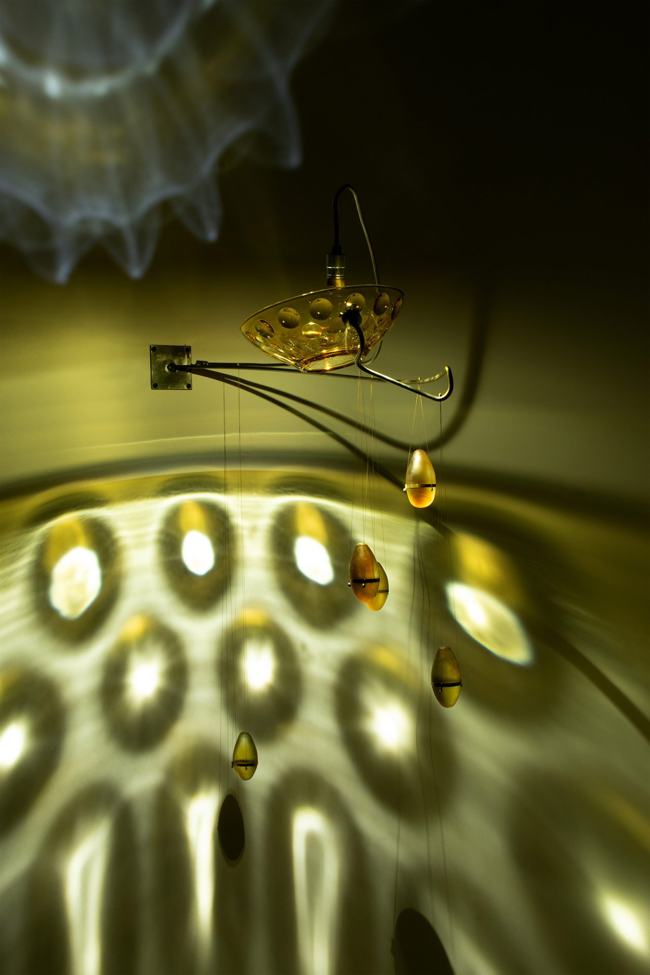 Wall-mounted fixture made of bent steel, brass accents, a yellow vintage coin dot glass bowl, and five yellow glass fishing weights. LED-lit, it casts yellow light in repeating circles.