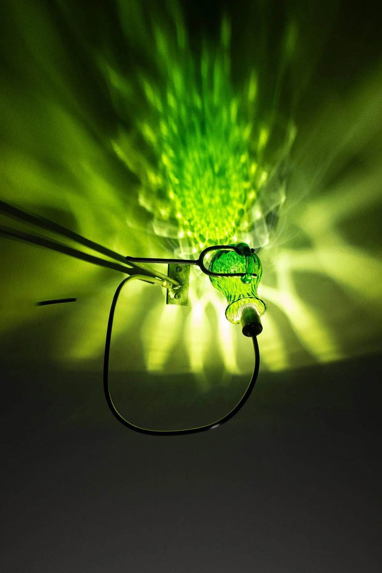 Wall-mounted fixture made of bent steel, brass accents, and a green vintage Empoli glass pitcher. LED-lit, it casts green light in a mandala-like pattern.