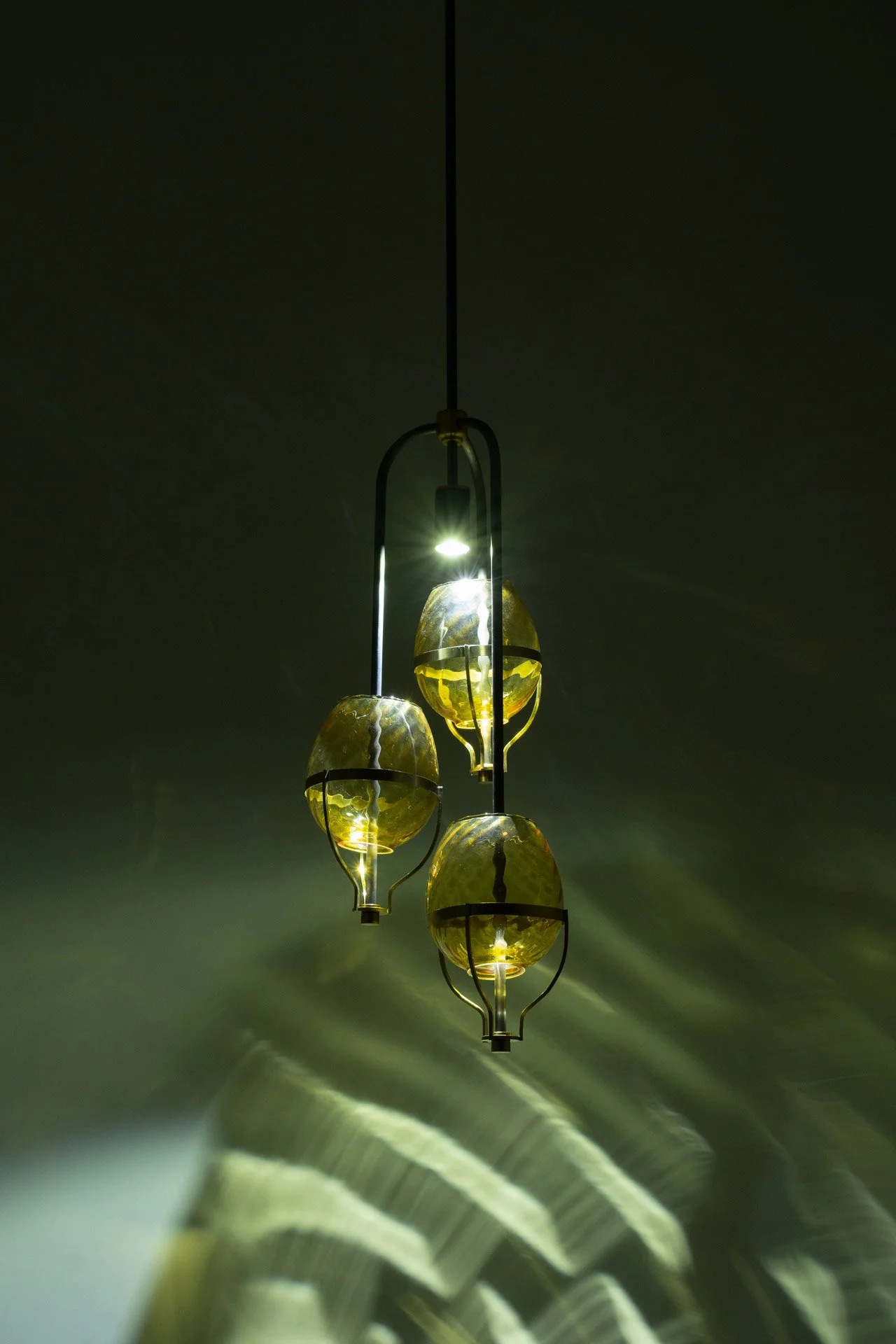 Ceiling pendant fixture made of bent steel piping, brass accents, and three yellow vintage swirled glass lampshades. LED-lit, it casts three distinct circles of swirled yellow light.
