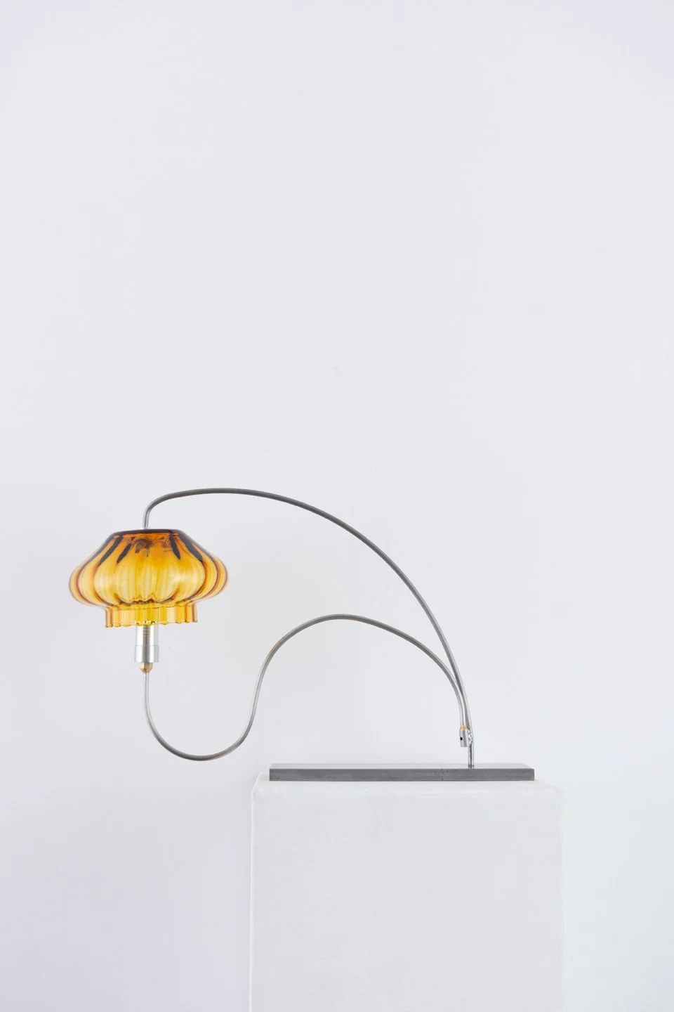 A modern table lamp with a curved metal arm and a yellow decorative lampshade, displayed on a white pedestal against a plain white background.