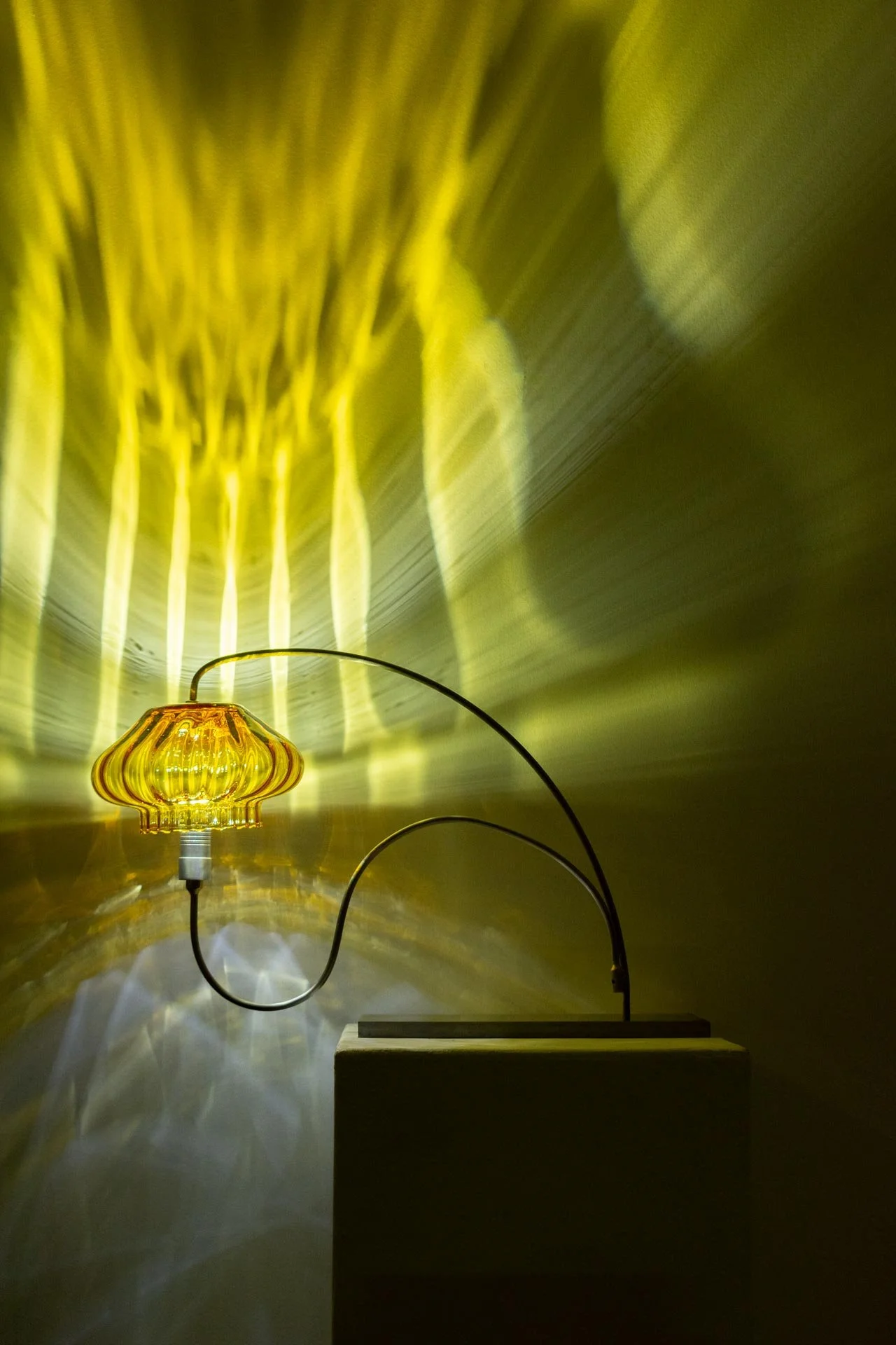 A yellow glass table lamp on a wooden stand with a curved black metal arm, casting intricate yellow light patterns onto the wall behind it.