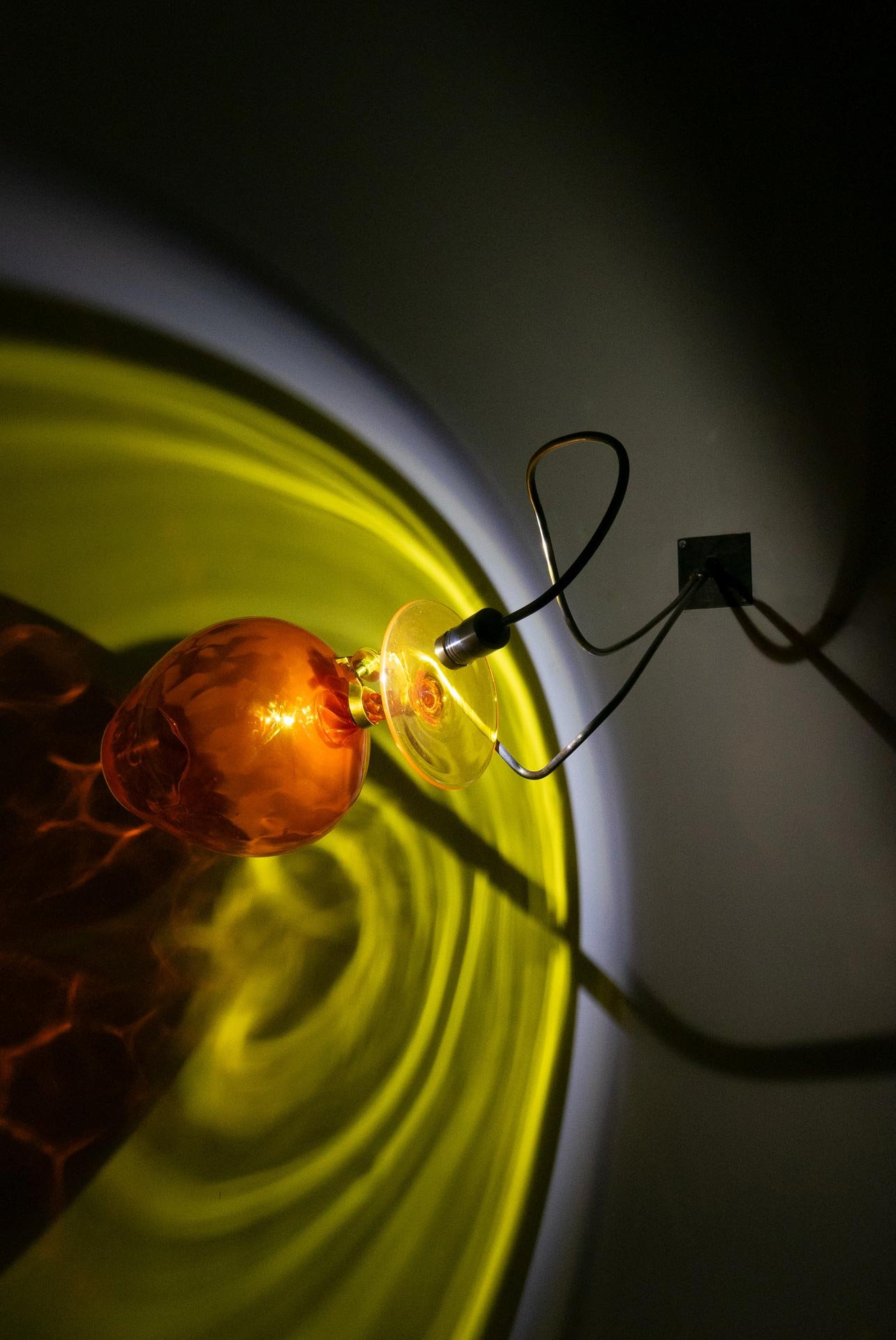 Wall-mounted fixture made of bent steel, brass accents, and an orange vintage glass goblet with a yellow base. LED-lit, layered orange, yellow, and blue light.