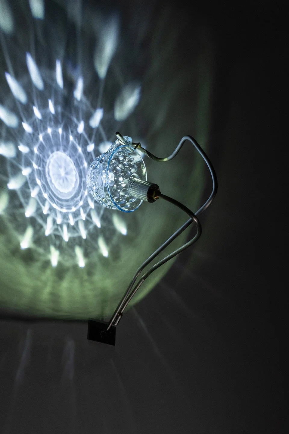 A glass lamp with a pointed shape, attached to a metallic stand, projecting intricate light patterns onto the wall.
