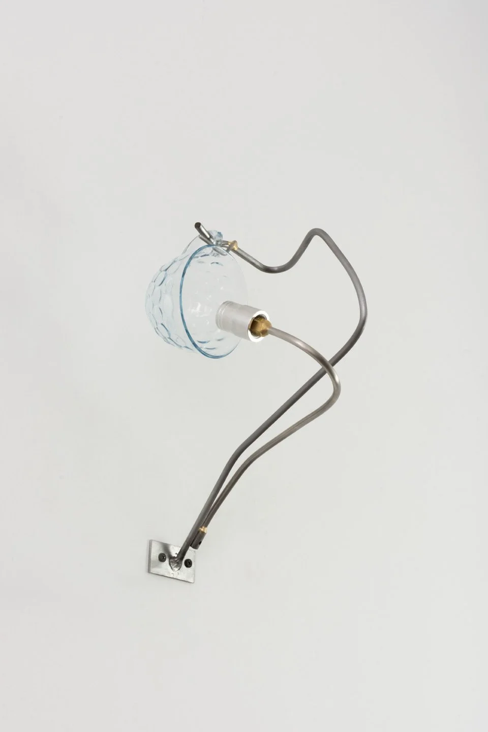 An abstract art sculpture resembling a bird made from metal, glass, and a light bulb, mounted on a white wall.