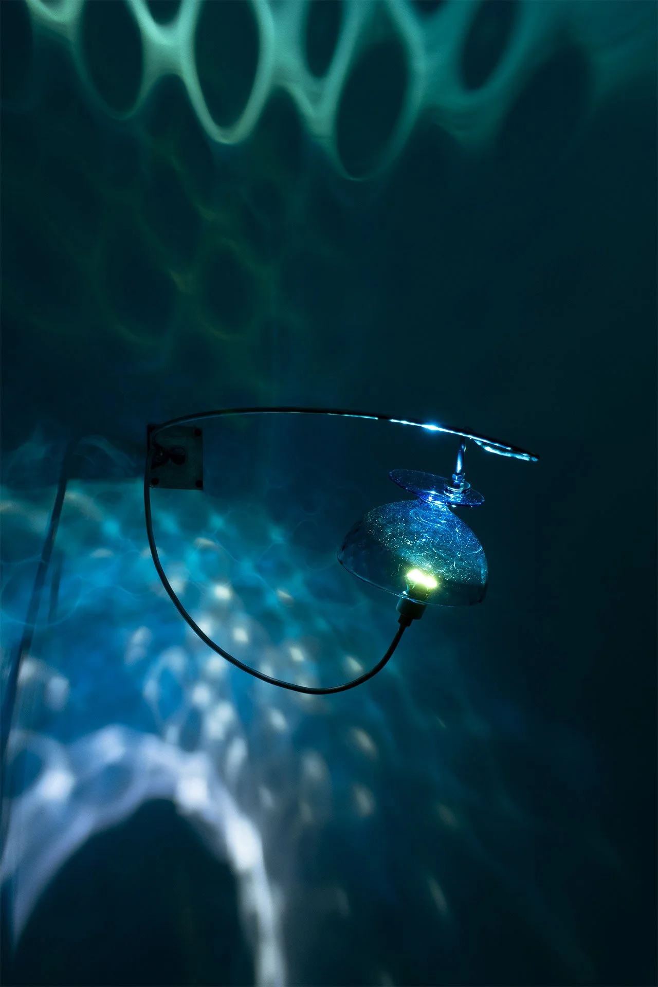 Wall-mounted fixture made of bent steel, brass accents, and a blue vintage glass tea cup. LED-lit, it casts blue light with a water-like pattern.
