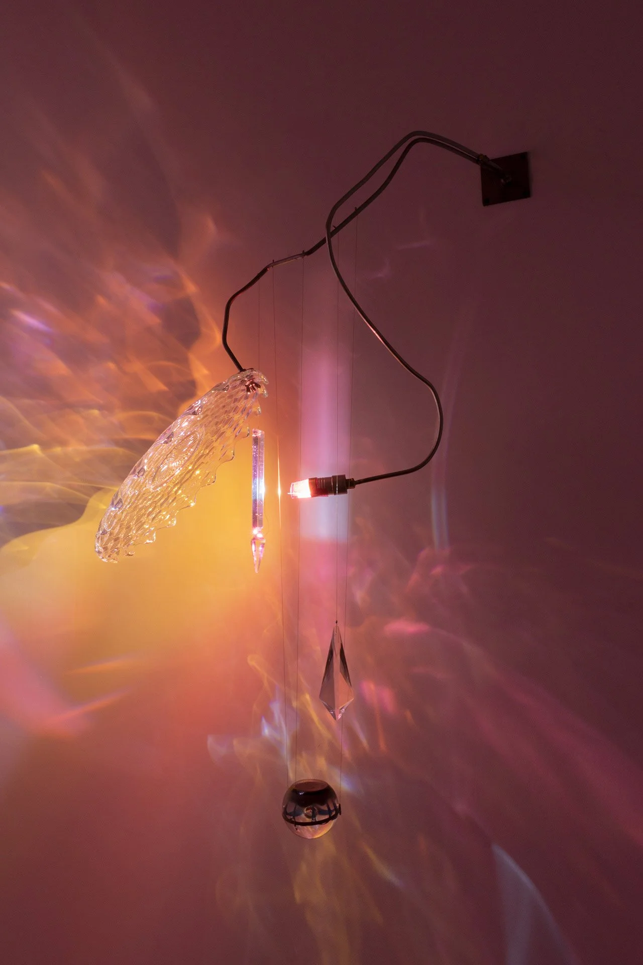 Wall-mounted fixture made of bent steel, brass accents, and vintage glassware: a clear Fostoria platter, two crystals, and a clear/blue spherical paperweight. LED-lit, it casts orange and pink light.