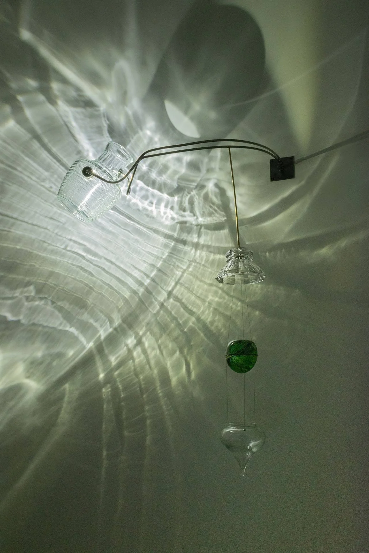 Wall-mounted fixture made of bent steel, brass accents, and vintage glassware: clear ribbed milk pitcher, lampshade, onion drop ornament, and green paperweight. LED-lit, it casts white ribbed light.