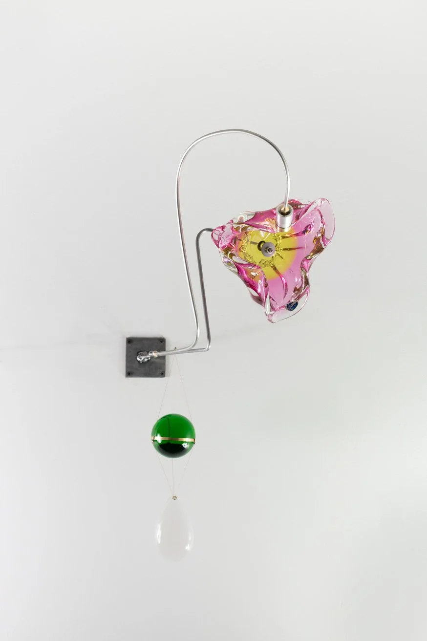 Modern art sculpture featuring a glass pink and yellow flower, a green and black sphere, and a glass teardrop, mounted on a white wall.