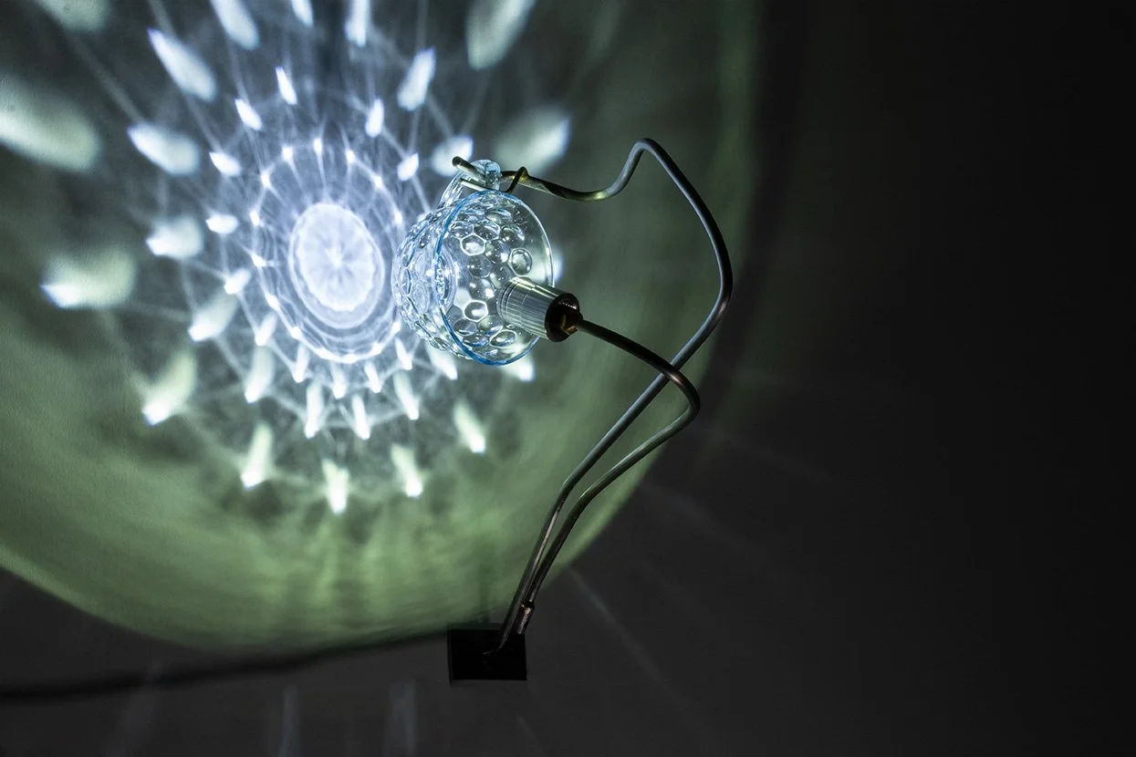 Wall-mounted fixture made of bent steel, brass accents, and a clear vintage coin-dot glass tea cup. LED-lit, it casts white light in a mandala-like pattern.